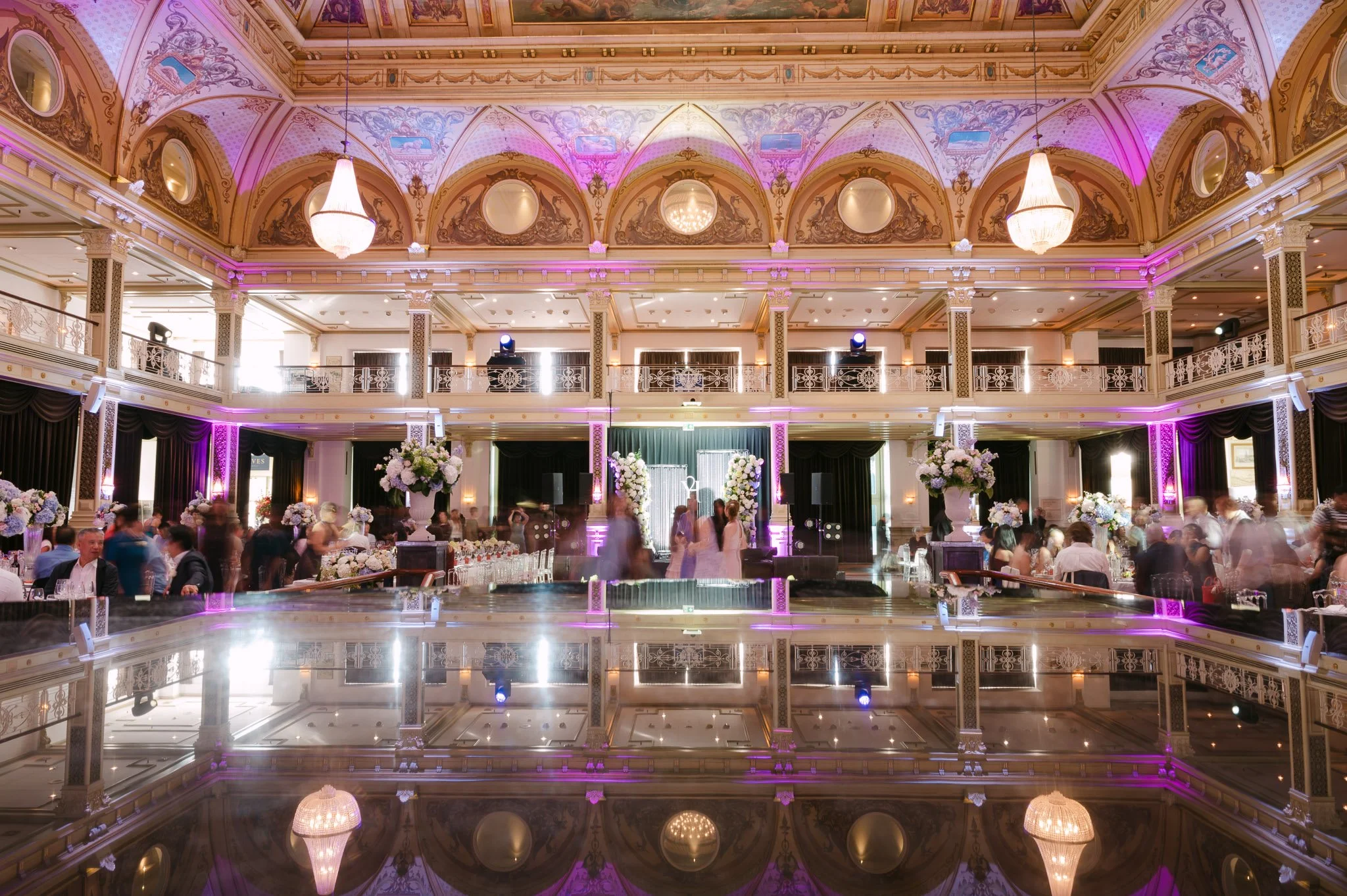 Elegant ballroom wedding reception with ornate ceiling, chandeliers, floral centerpieces, purple lighting, and guests.