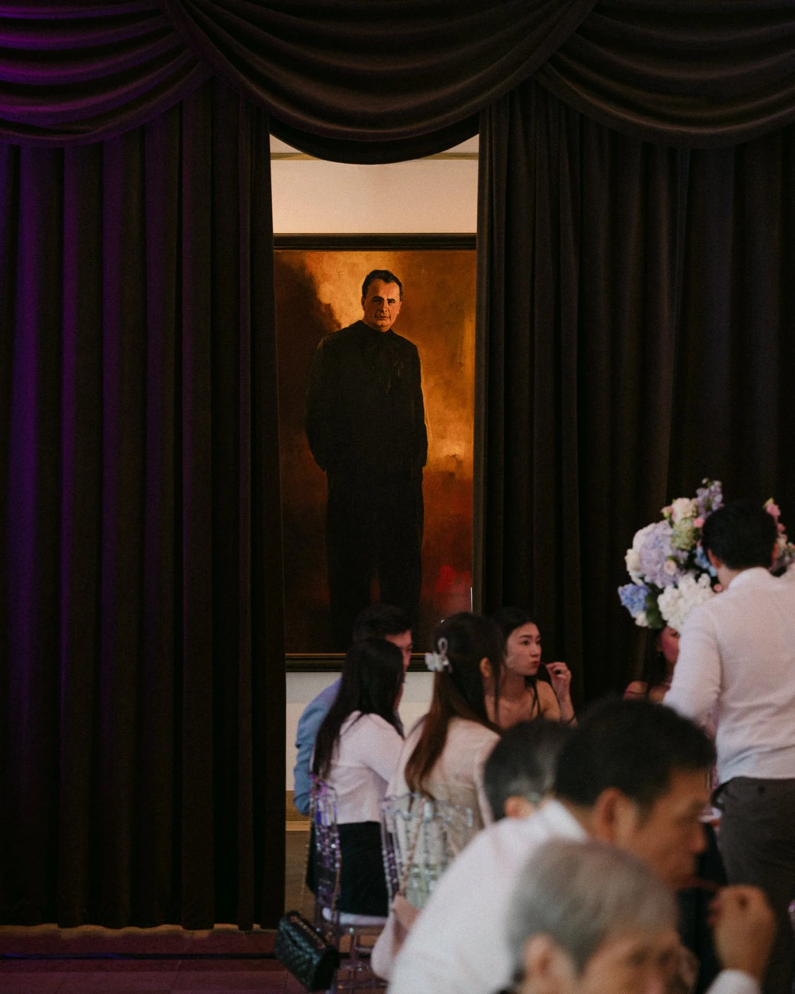 Guests gather beneath black theater curtains beside a portrait of a man in a dark suit and a large floral arrangement.
