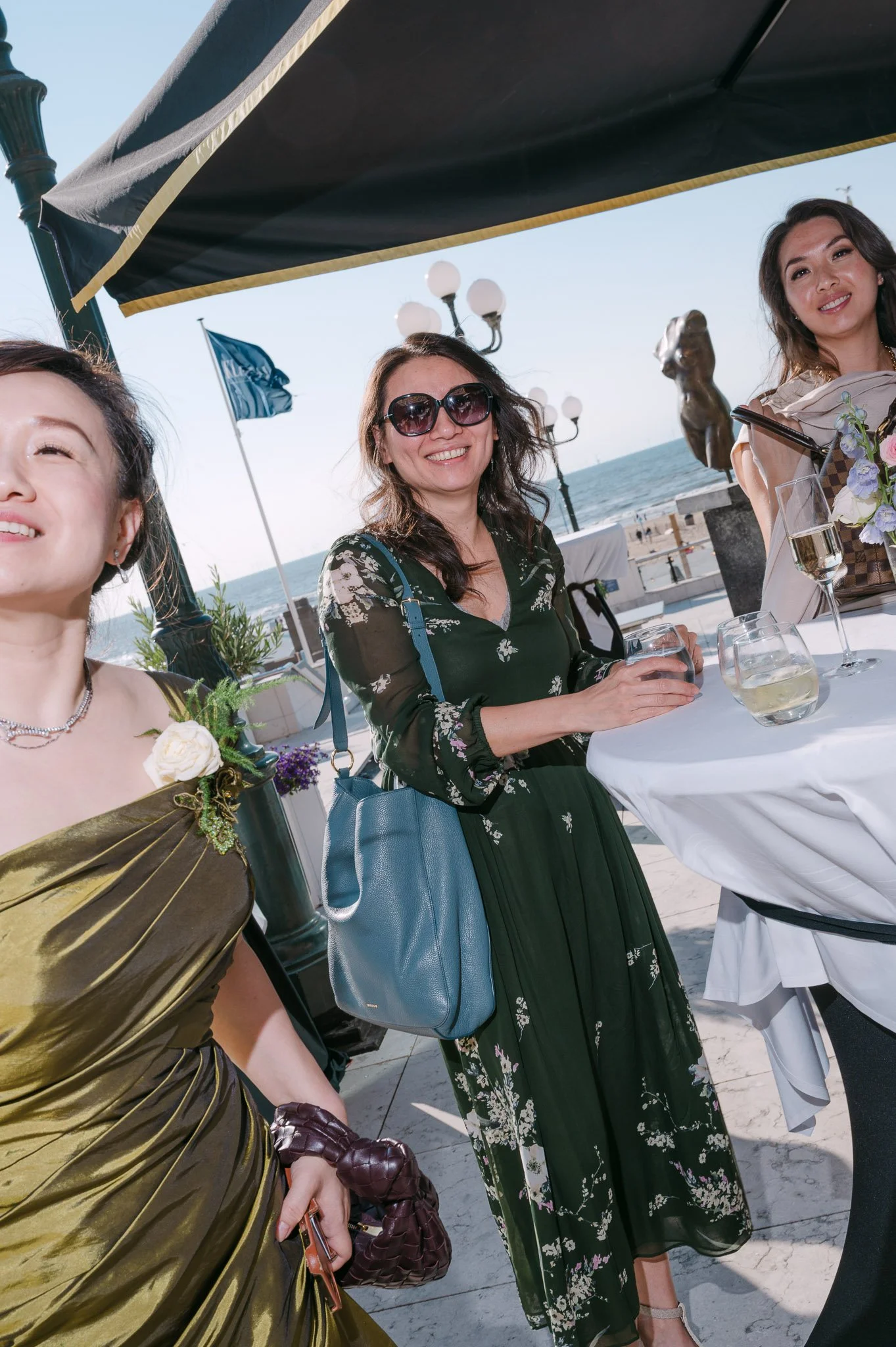 Three women enjoying drinks at an elegant seaside restaurant beneath a patio umbrella.