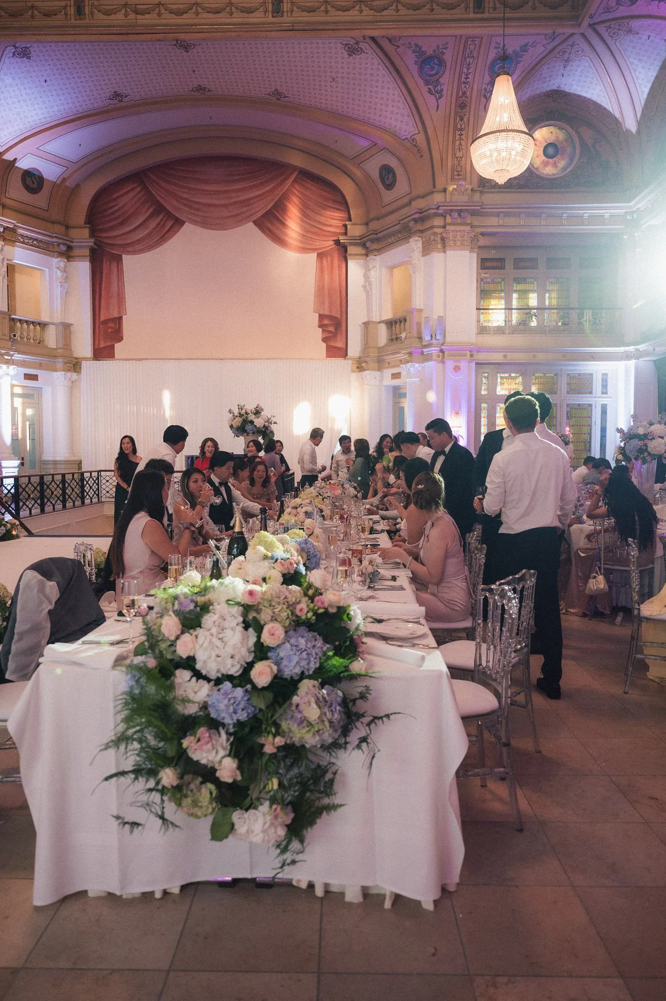 Elegant wedding reception with guests dining at flower-adorned tables beneath ornate chandeliers in a historic ballroom