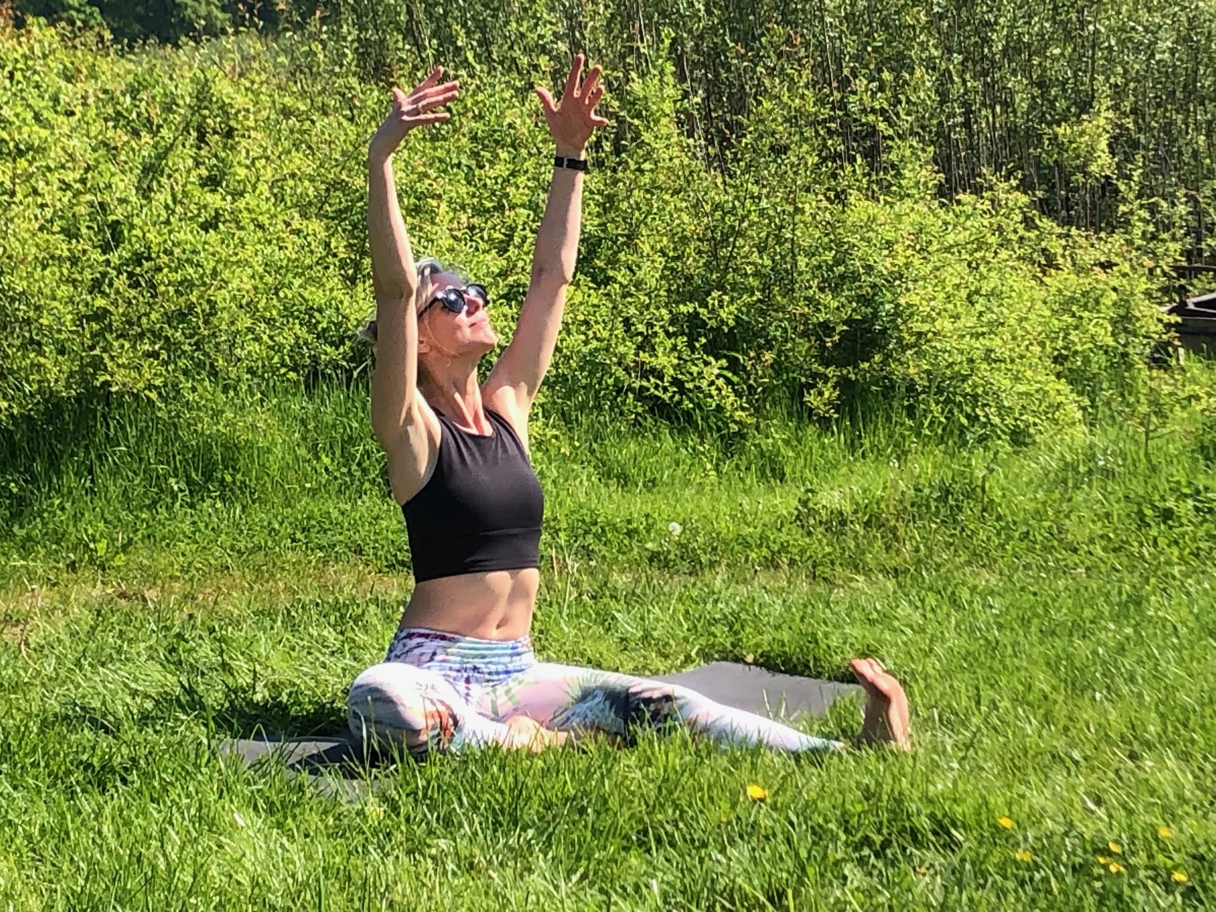 Woman practicing yoga outdoors on a mat in a grassy field, seated in a wide-leg pose with arms raised.