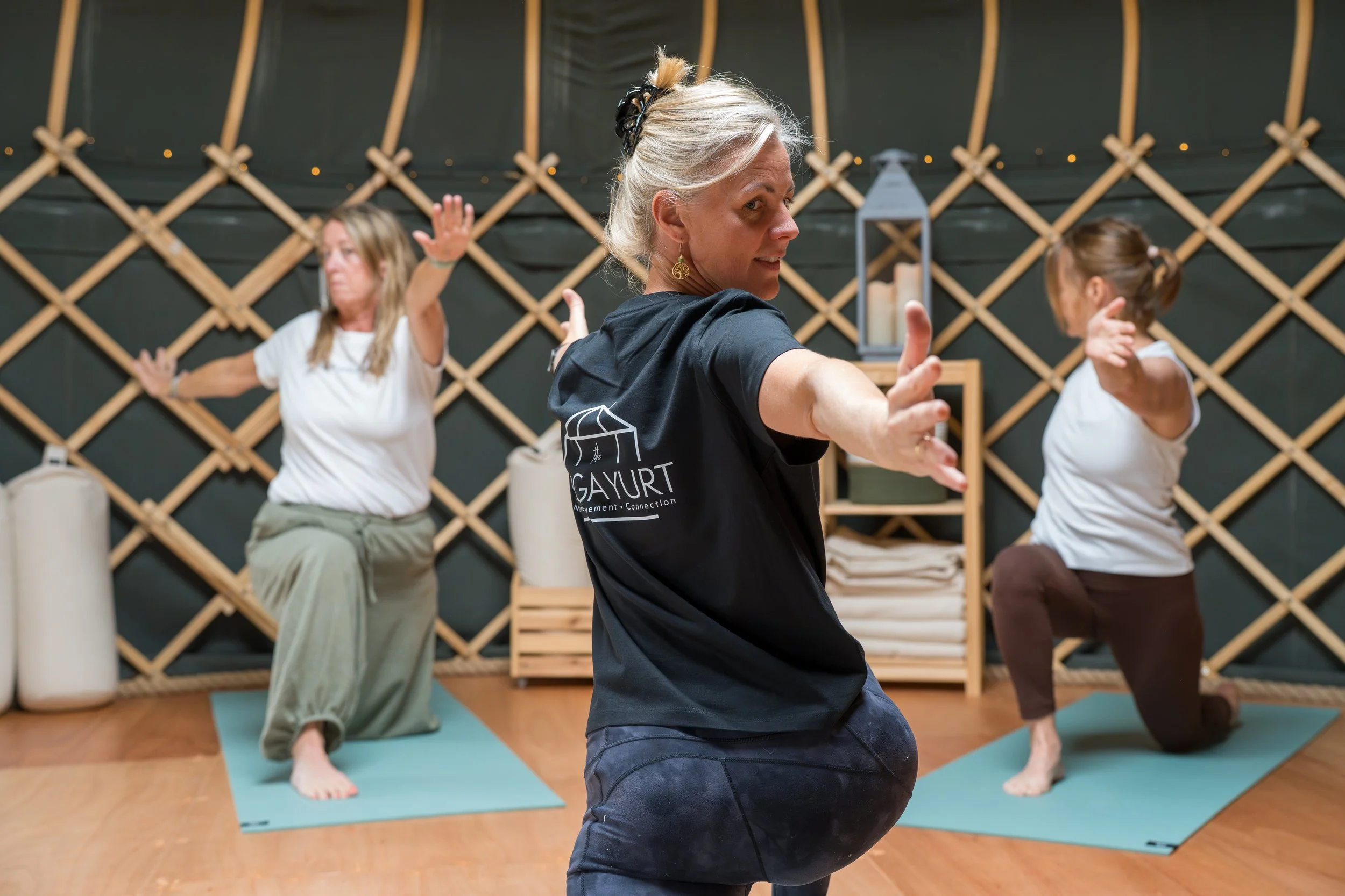 Three women practicing seated yoga poses on mats in a studio, led by an instructor in the foreground