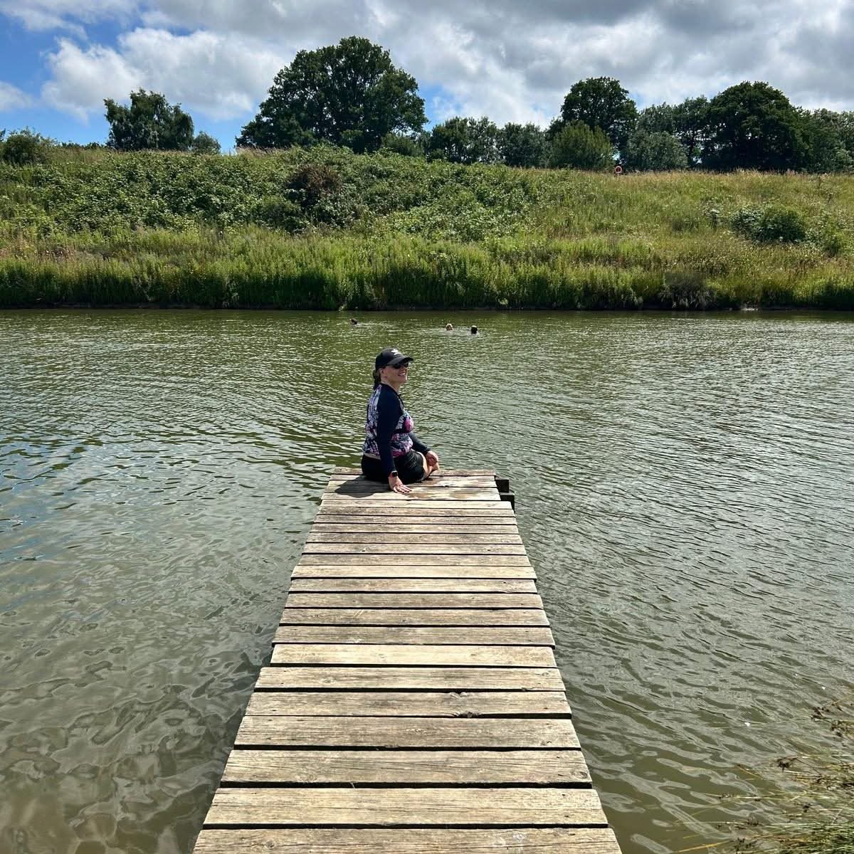 Woman sitting on the end of a wooden dock extending into a calm lake, with grassy banks and trees under a partly cloudy sky.