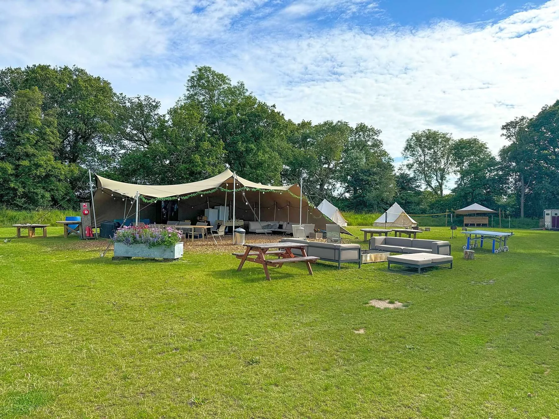 Outdoor glamping campsite with canvas tents, lounge seating, picnic tables, table tennis, and trees on a sunny lawn