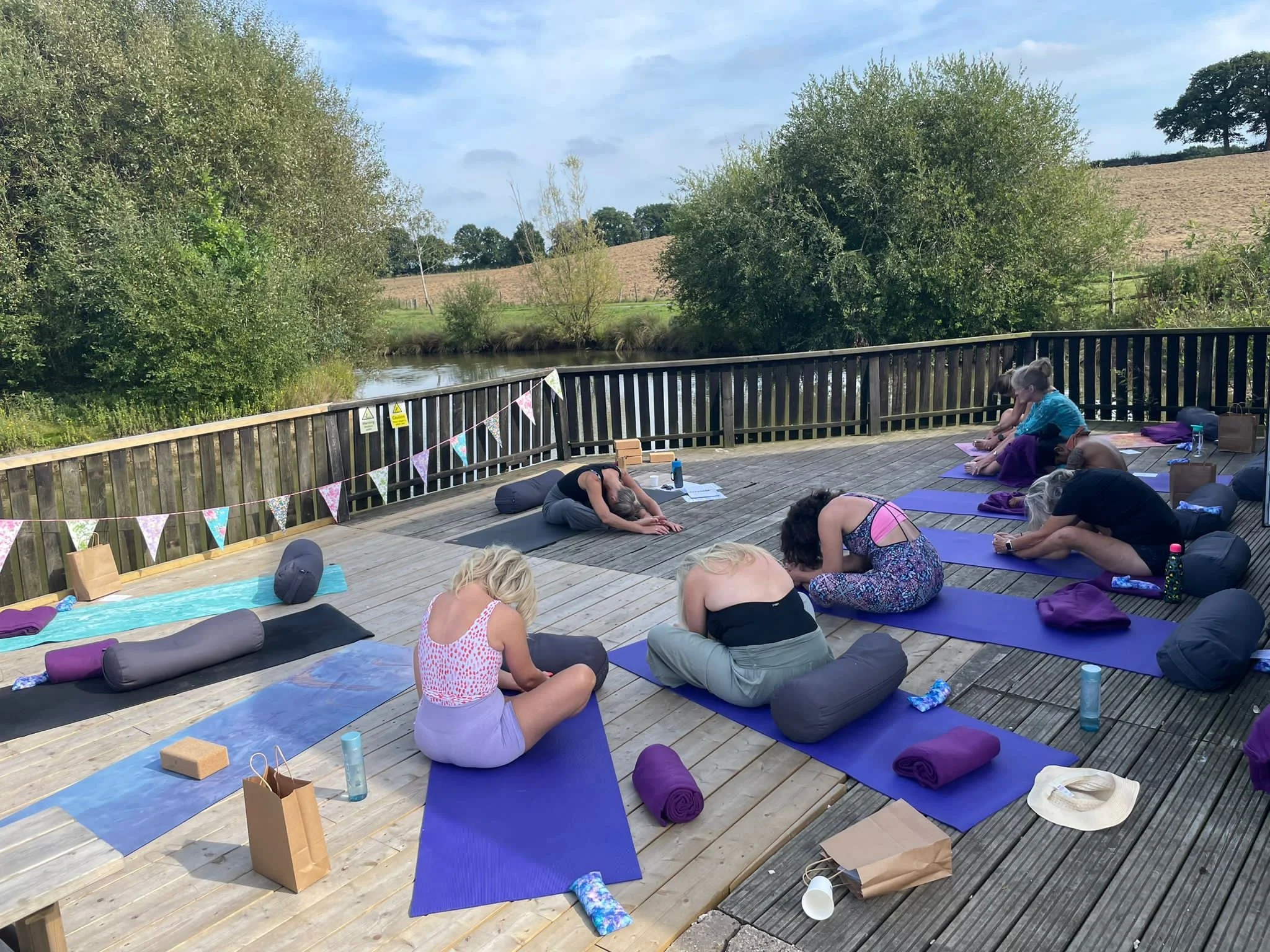 Outdoor yoga class on a wooden deck beside a river, with participants practicing stretches on colorful mats and rolling countryside behind them.