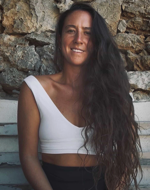 Smiling woman with long dark hair wearing a white crop top, seated against a rustic stone wall