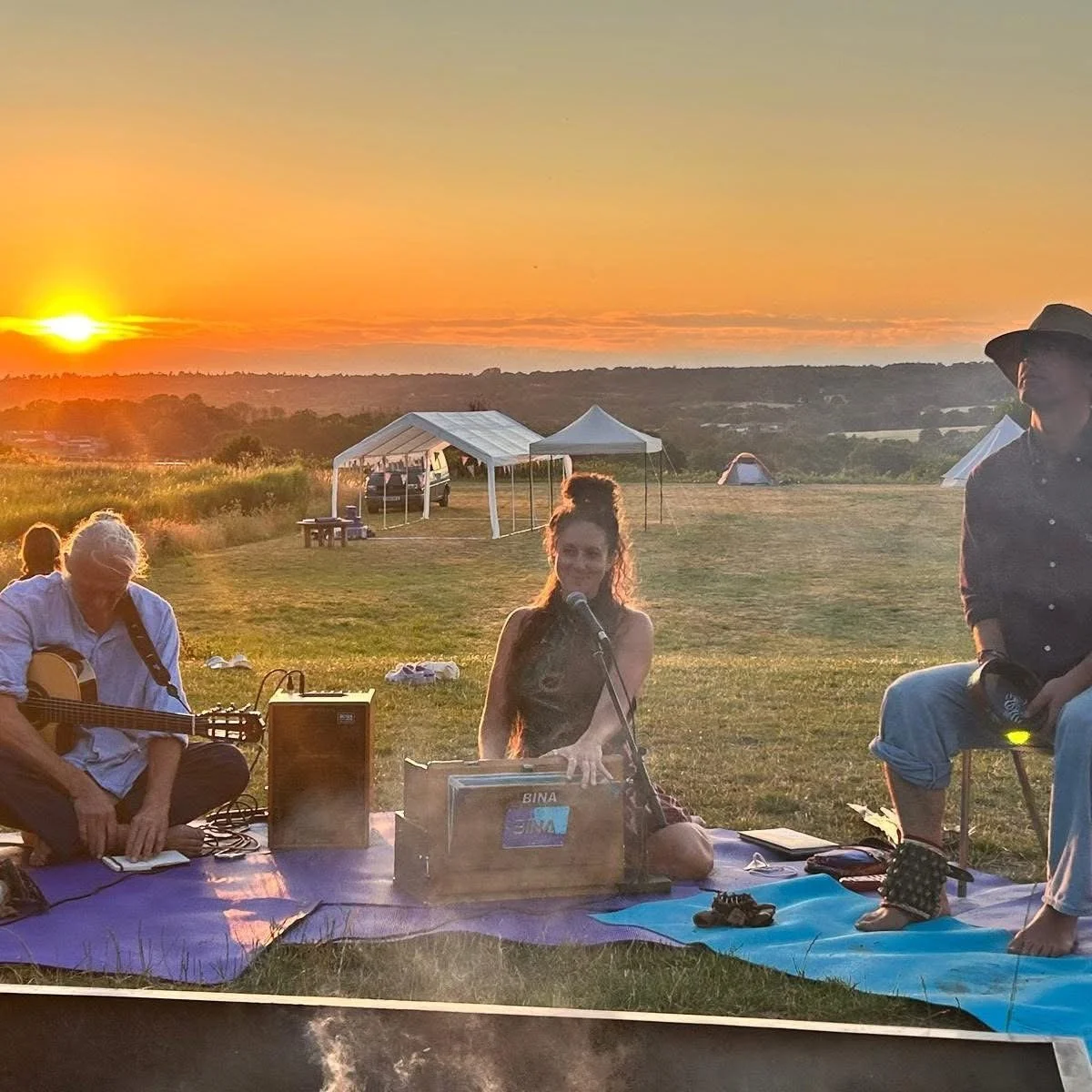 Musicians perform outdoors at sunset on a grassy hillside, with a guitar, harmonium, tents, and canopy tents in the background.