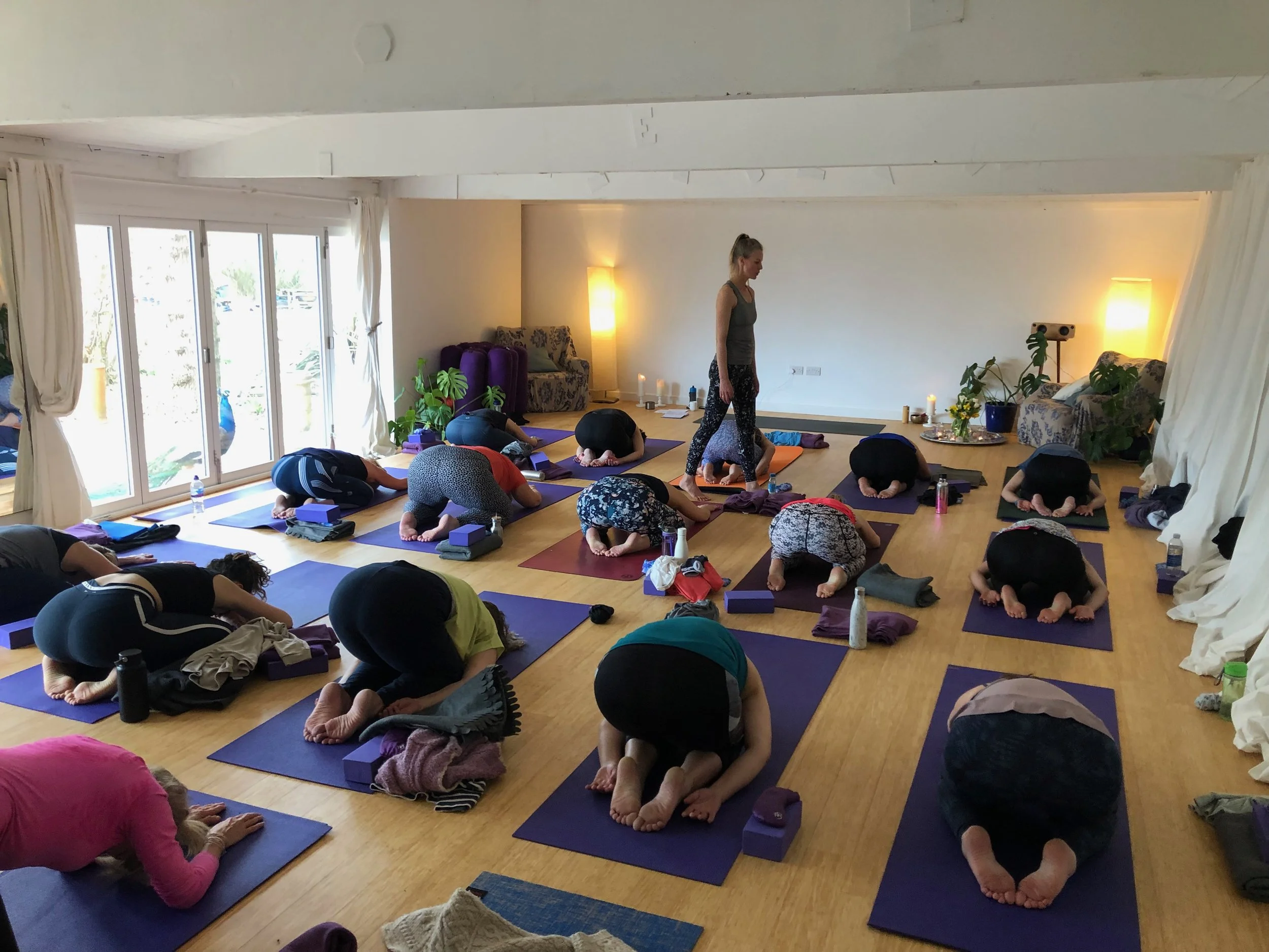 Yoga class in a bright studio with participants practicing child’s pose on mats while an instructor stands nearby.