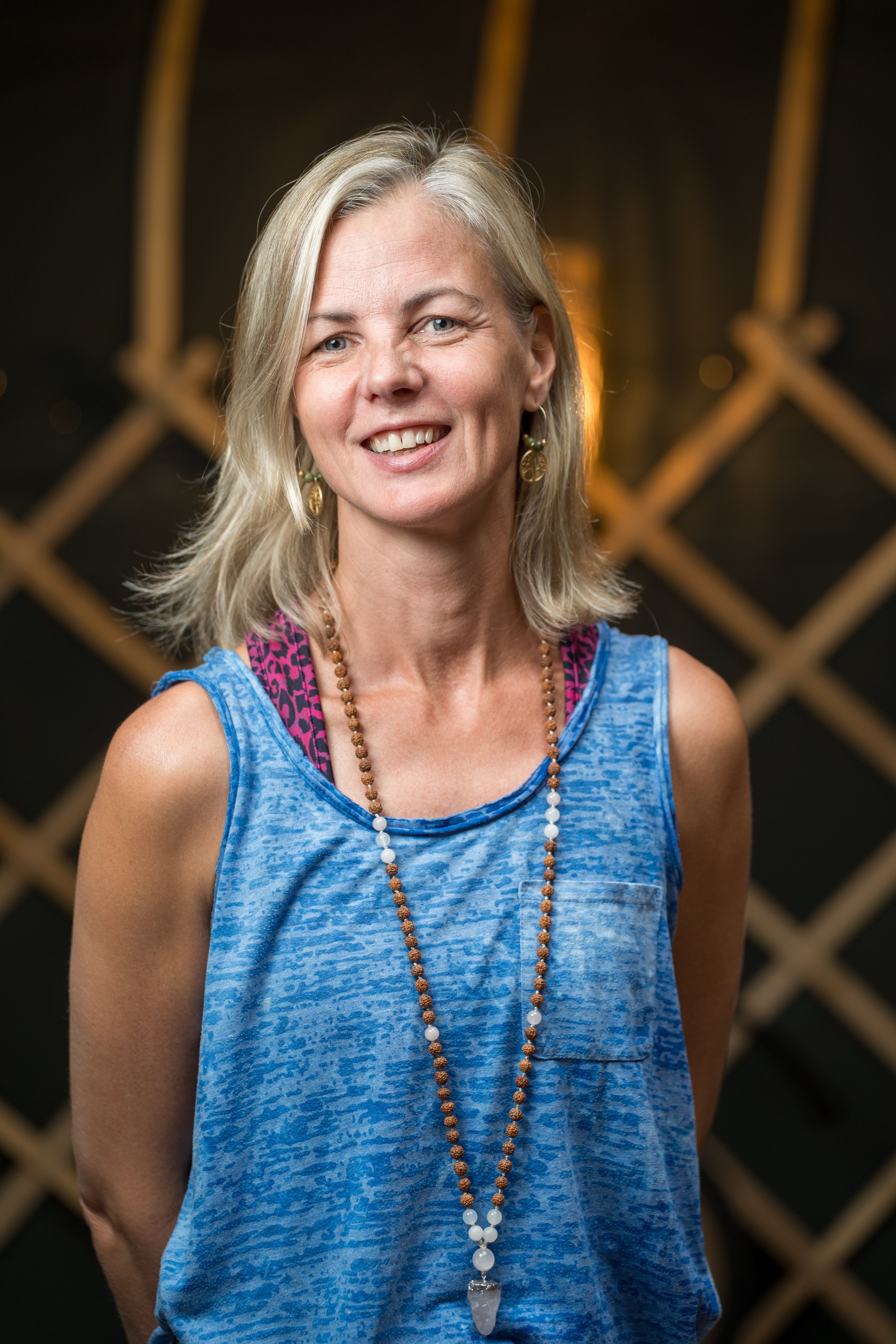 Smiling blonde woman wearing a blue sleeveless top and beaded necklace in a warmly lit setting