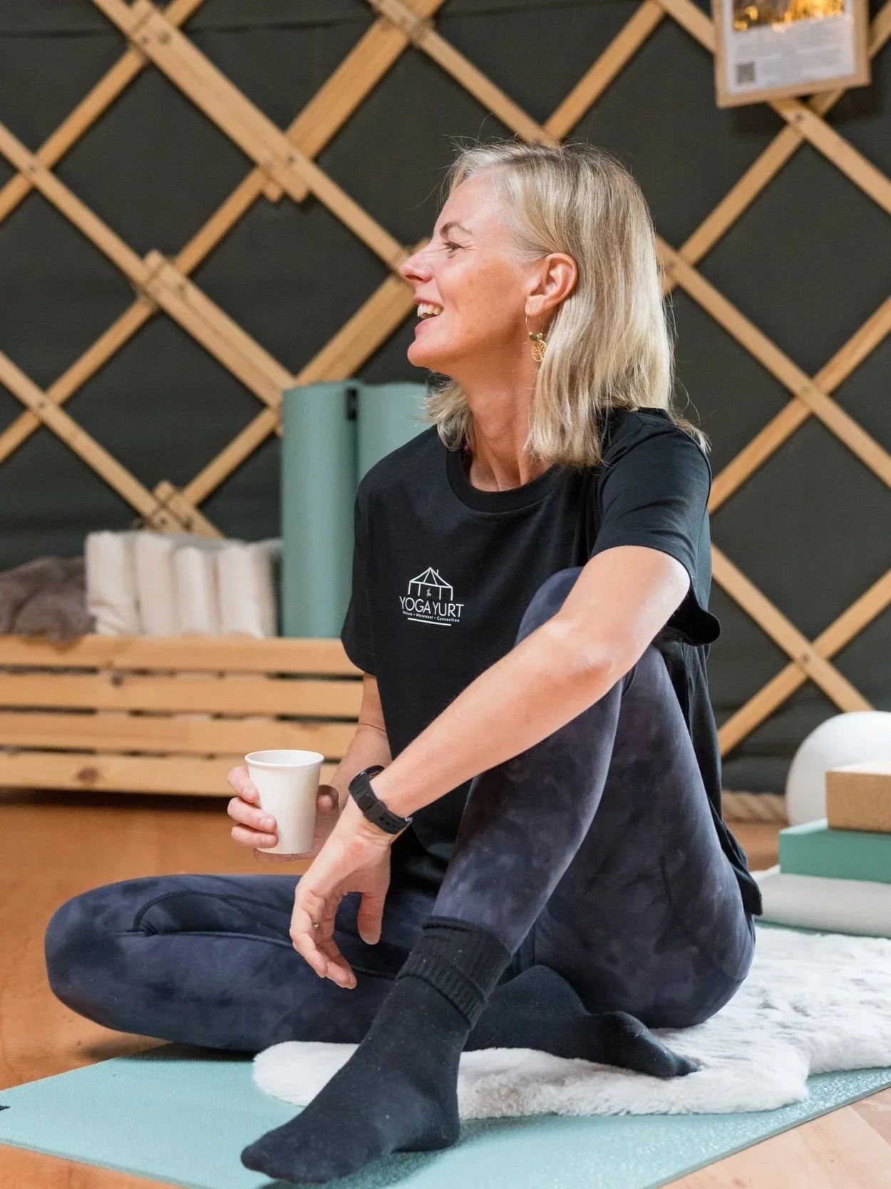 Smiling woman in a Yoga Yurt shirt sitting cross-legged on a yoga mat and holding a cup