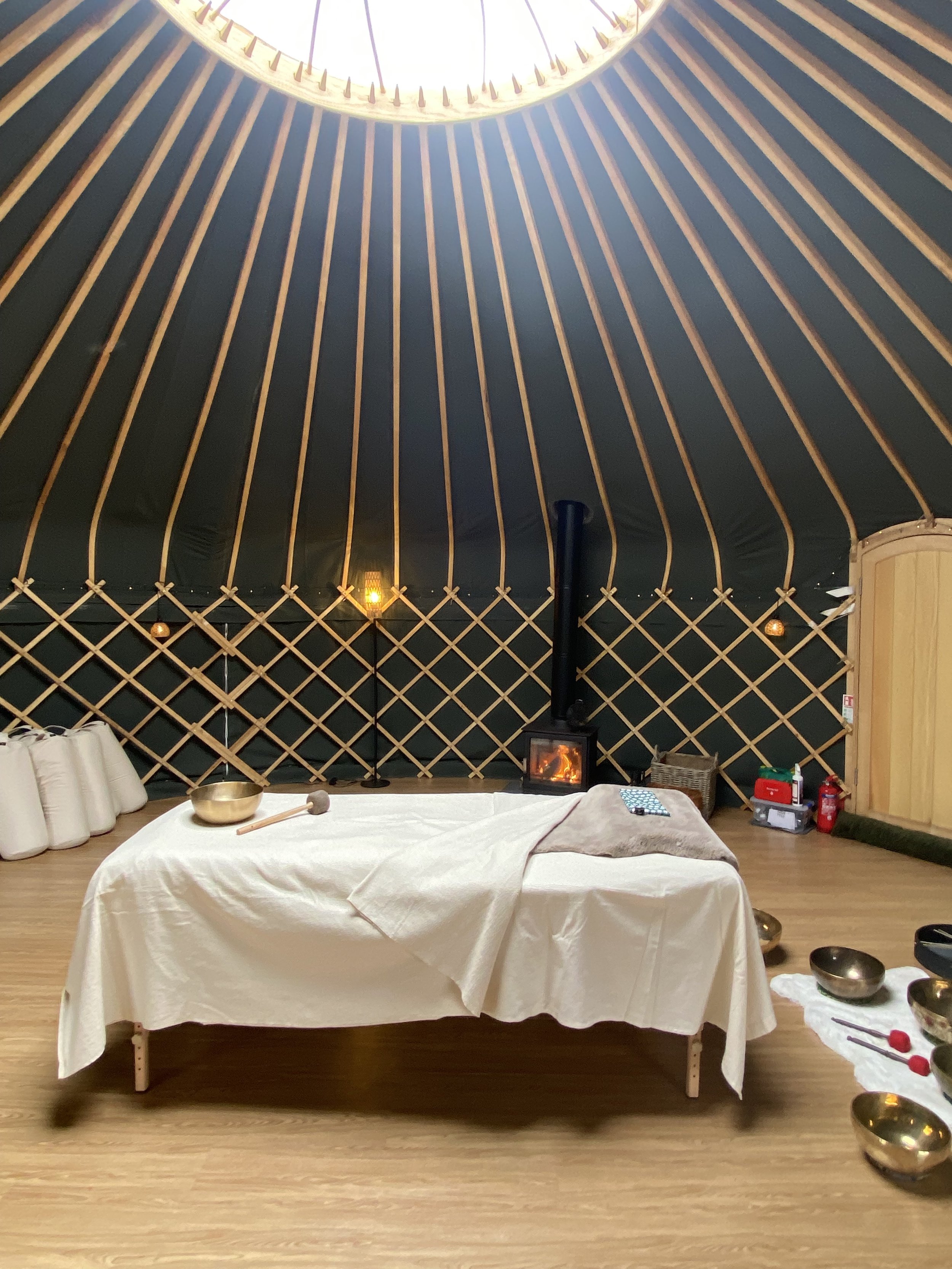 Interior of a yurt-style wellness room with a wood-burning stove, massage table, singing bowls, and wooden lattice walls.