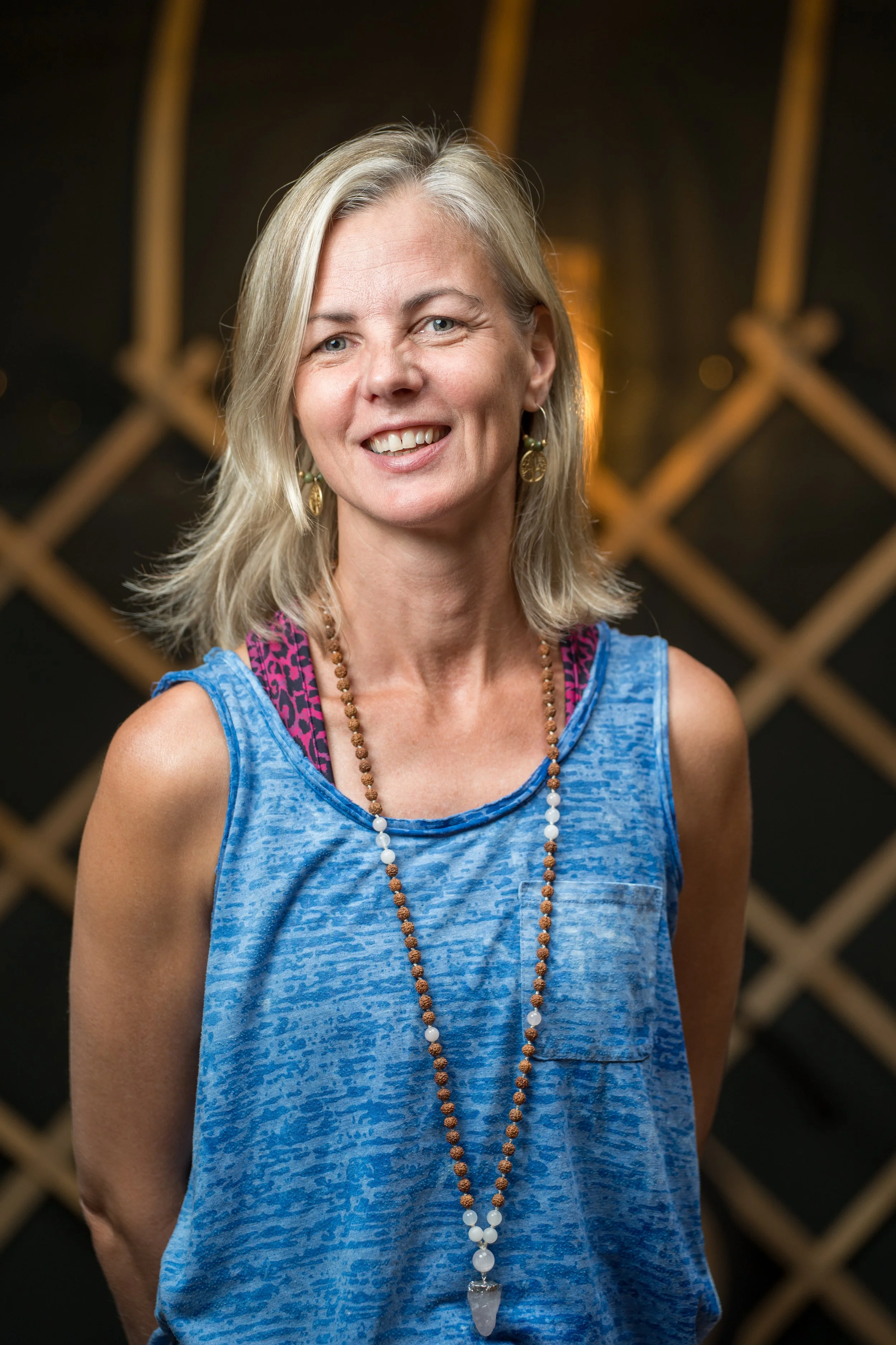 Smiling blonde woman wearing a blue sleeveless top and beaded necklace in front of a dark wooden background
