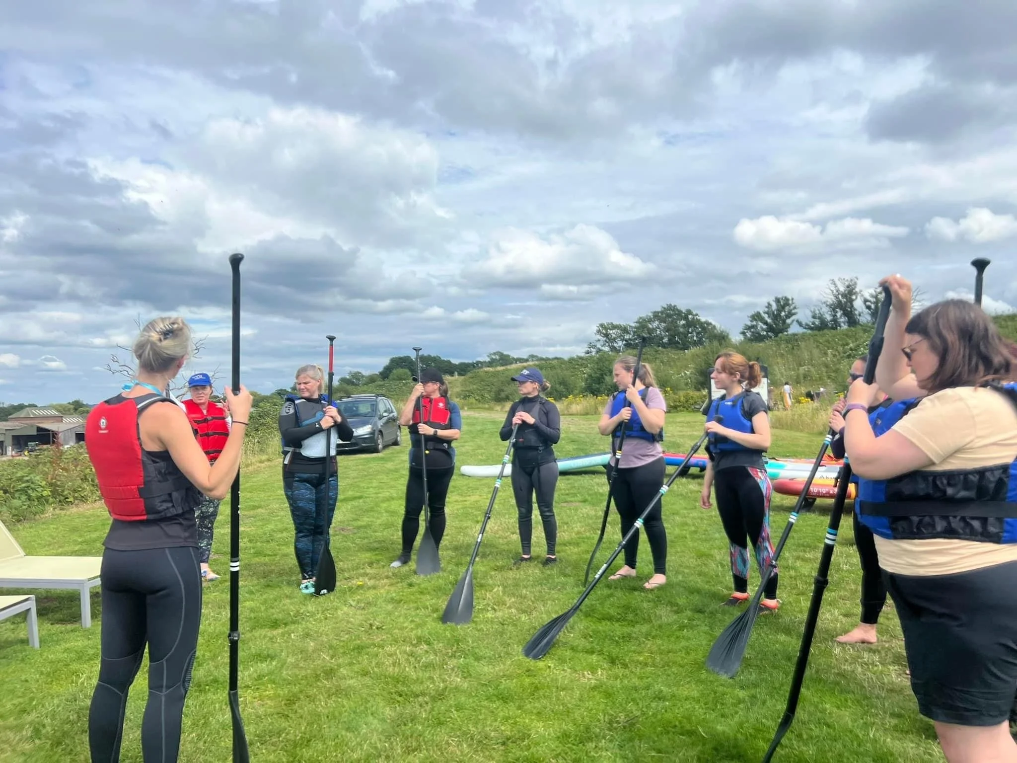 Group of people wearing life jackets and holding paddles during an outdoor paddleboarding lesson on a grassy shore