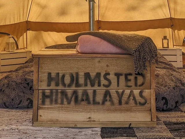 Rustic tent interior with a wooden crate labeled “HOLMSTED HIMALAYAS,” bedding, and a rolled blanket.