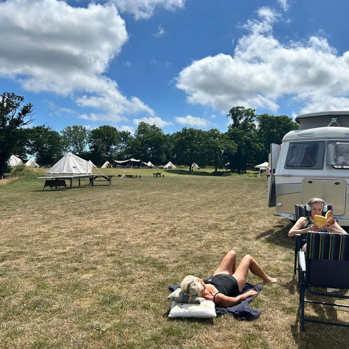 Sunny campground with bell tents, a caravan, and people relaxing on the grass beneath a blue sky with clouds