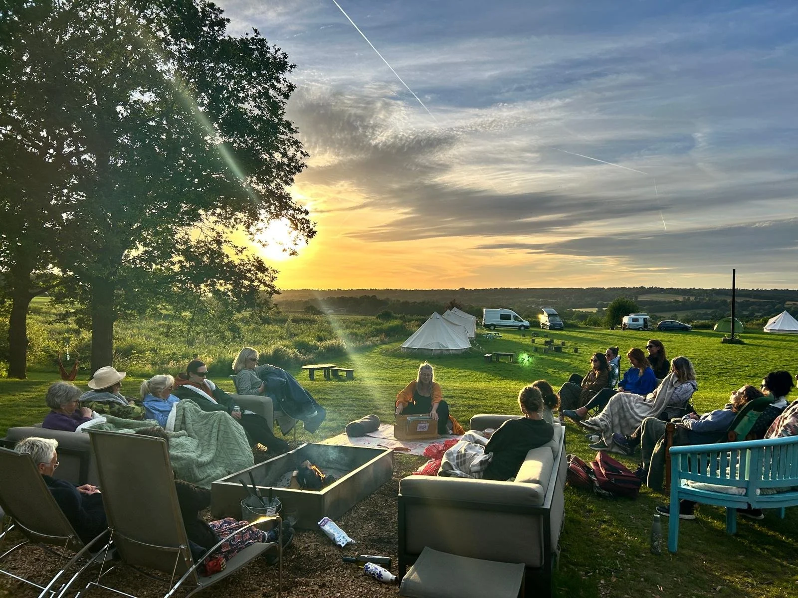 Group of people relaxing around an outdoor fire pit at sunset on a grassy campsite with tents, vans, and rolling countryside.