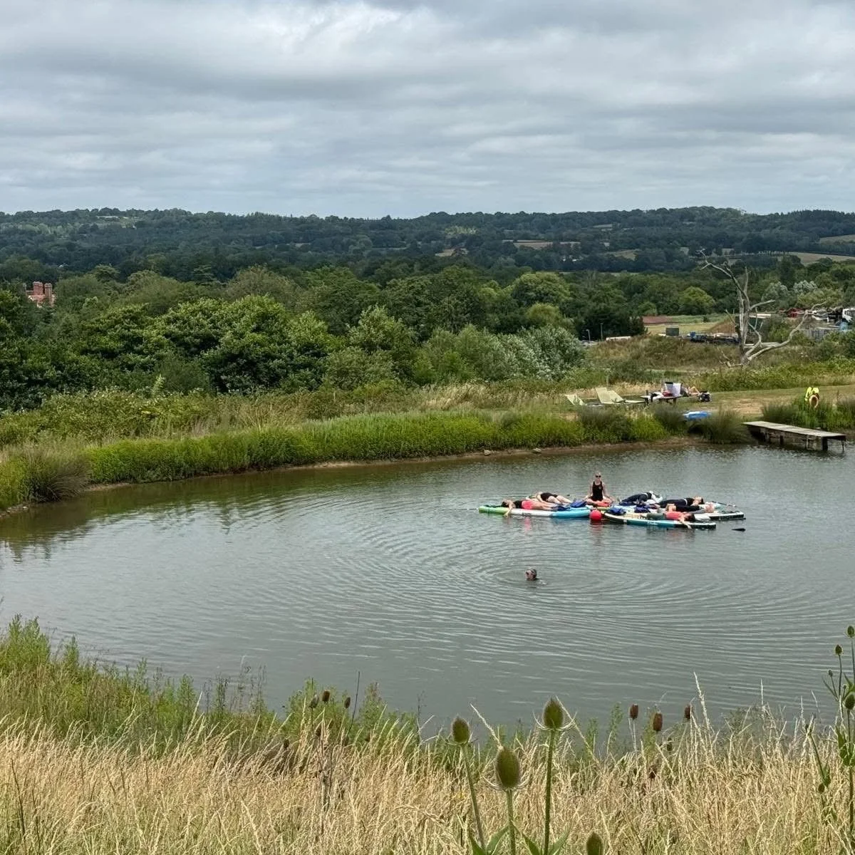 People relaxing on paddleboards in a calm lake surrounded by grassy fields and wooded hills under an overcast sky