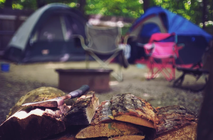 Camping scene with split firewood and hatchet in the foreground, tents and folding chairs in the background