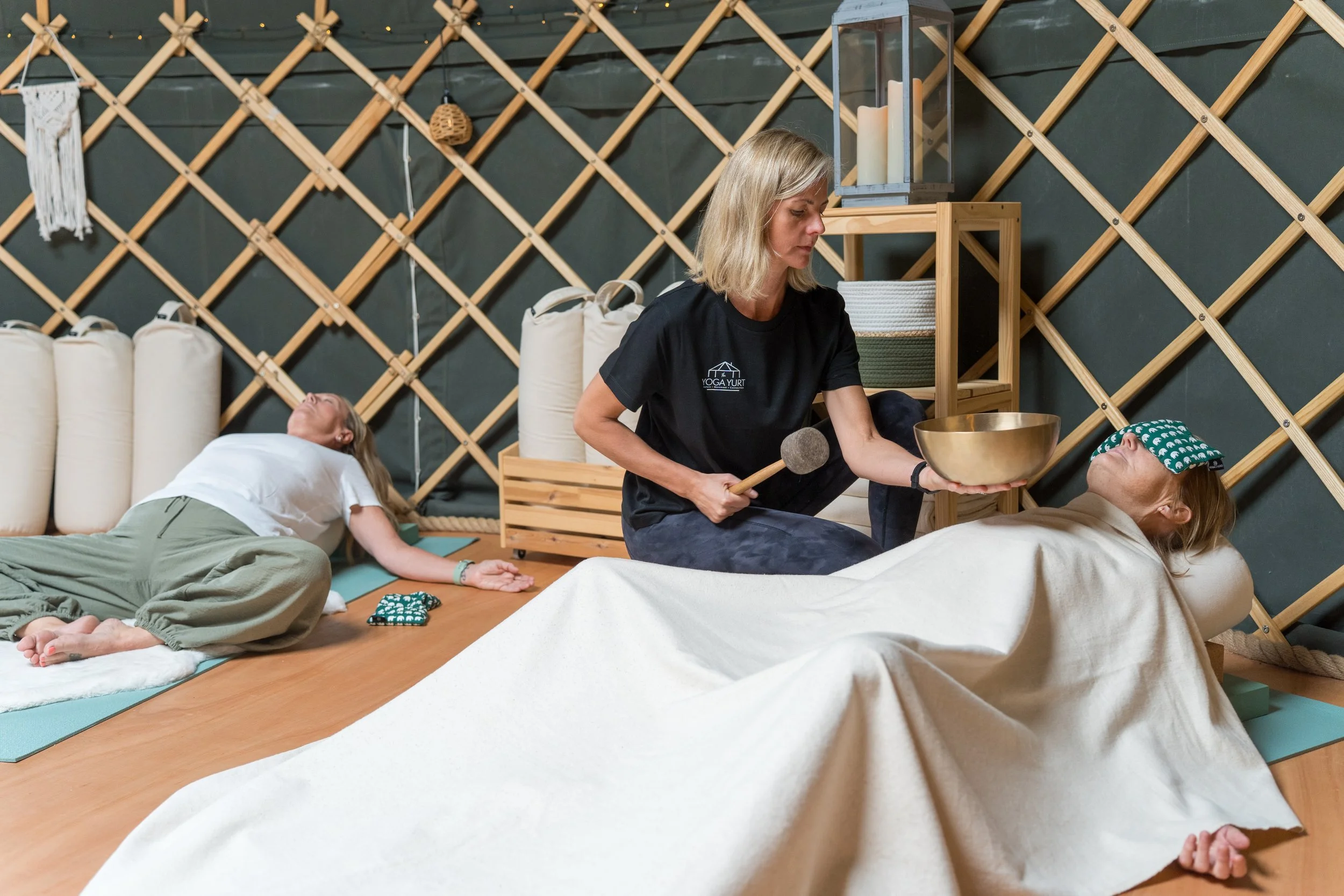 Sound bath instructor playing a singing bowl and mallet for two reclining participants in a cozy yoga studio.