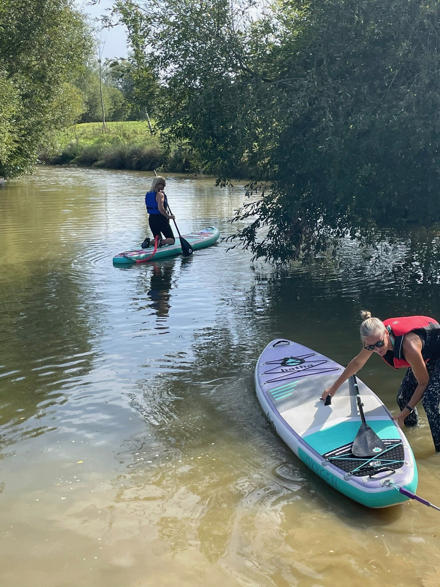 Two women paddleboarding on a tree-lined river, one wearing a life jacket and kneeling on a colorful paddleboard while another adjusts her board in the foreground.