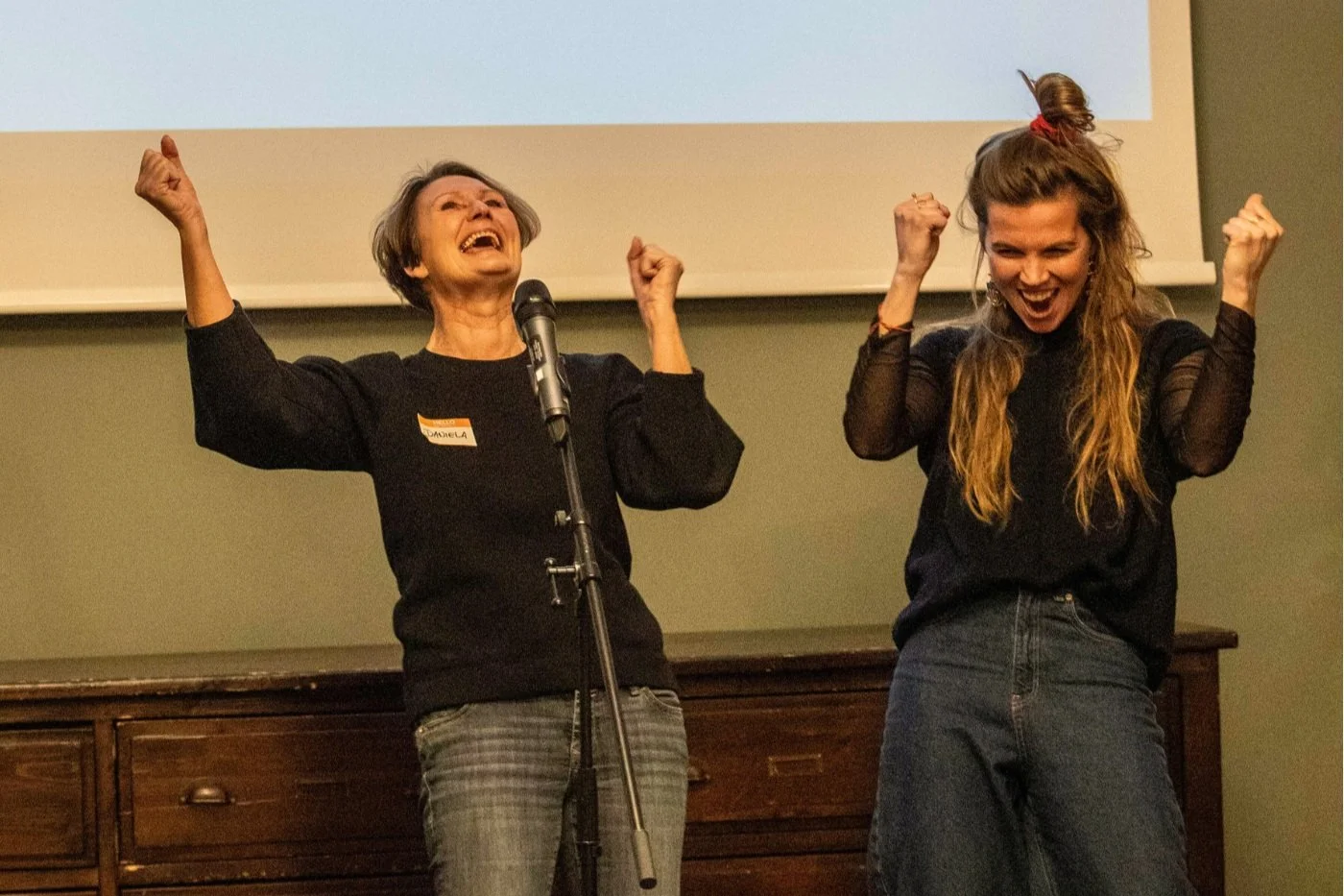 Two women, 1 coach and 1 coachee, cheering happy, behind microphone