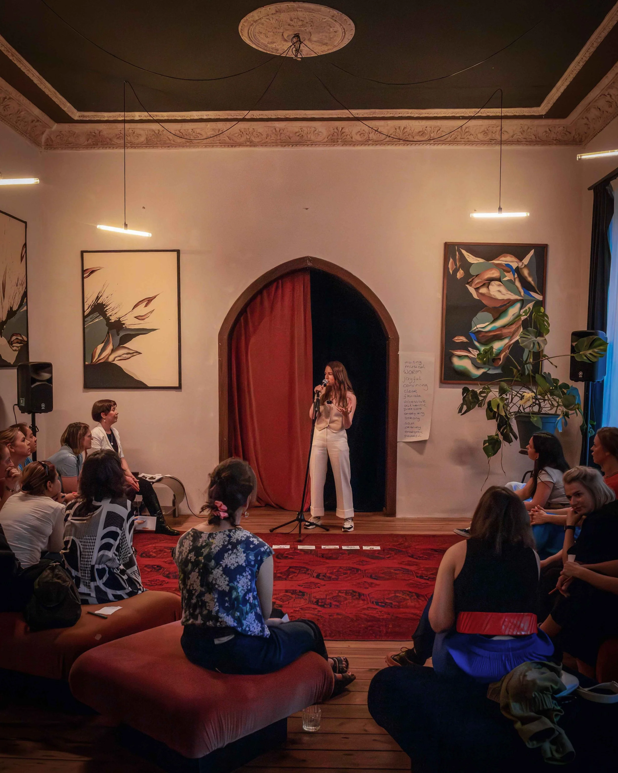 Colourful, creative room, with participants in workshop setting and 1 woman behind microphone