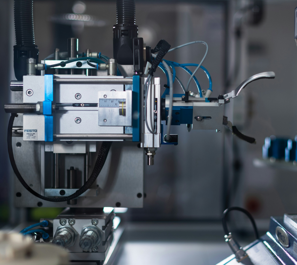 Close-up of a robotic automation device with metal components, wires, and pneumatic tubes in a laboratory or manufacturing setting.
