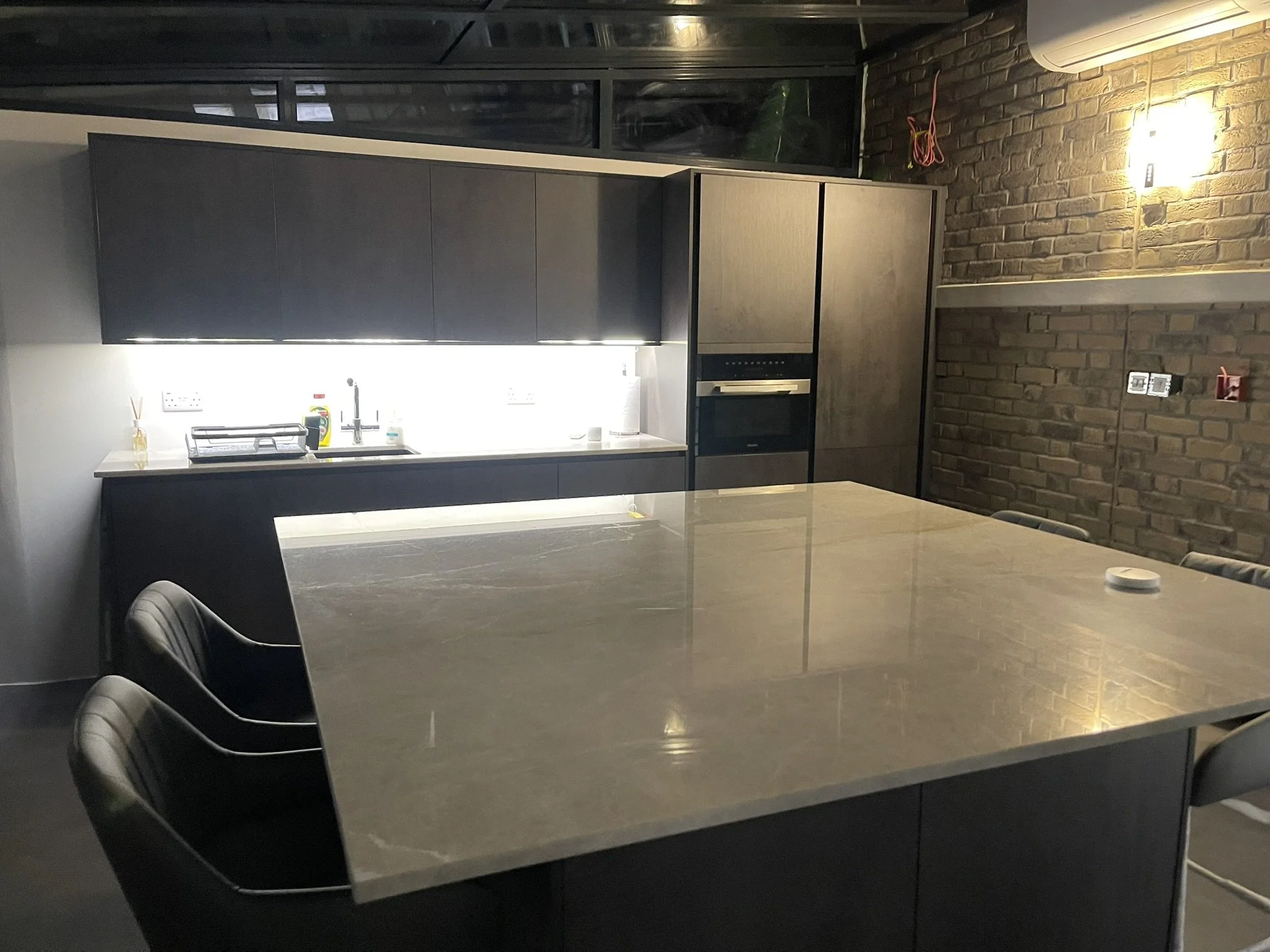 Modern dark kitchen with a large central island, black cabinets, stainless steel appliances, and exposed brick walls