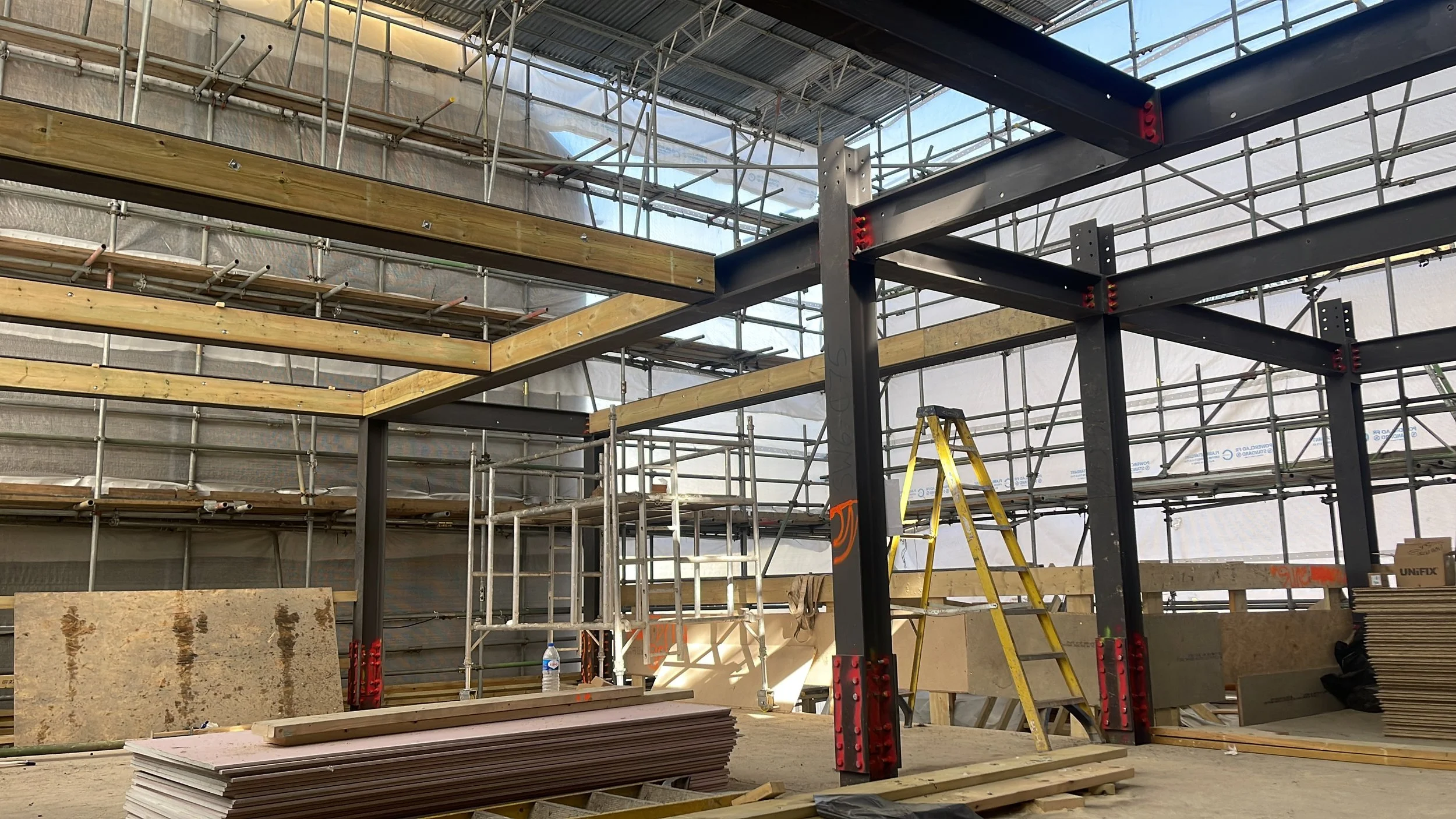 Steel-framed building under construction with scaffolding, wooden beams, plywood panels, and a yellow ladder
