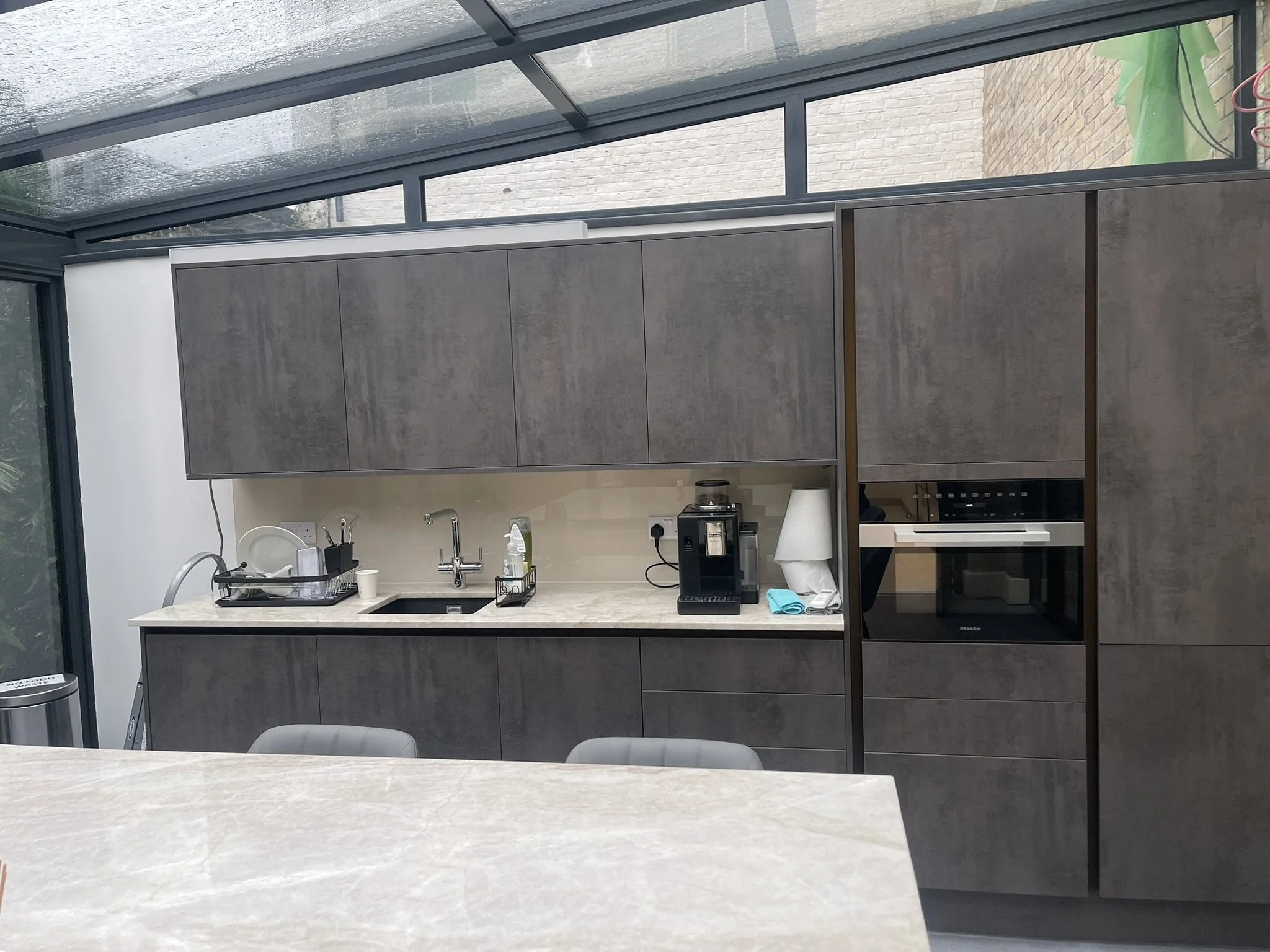 Modern dark-paneled kitchen with marble countertops, sink, dish rack, coffee maker, built-in oven, and glass conservatory roof