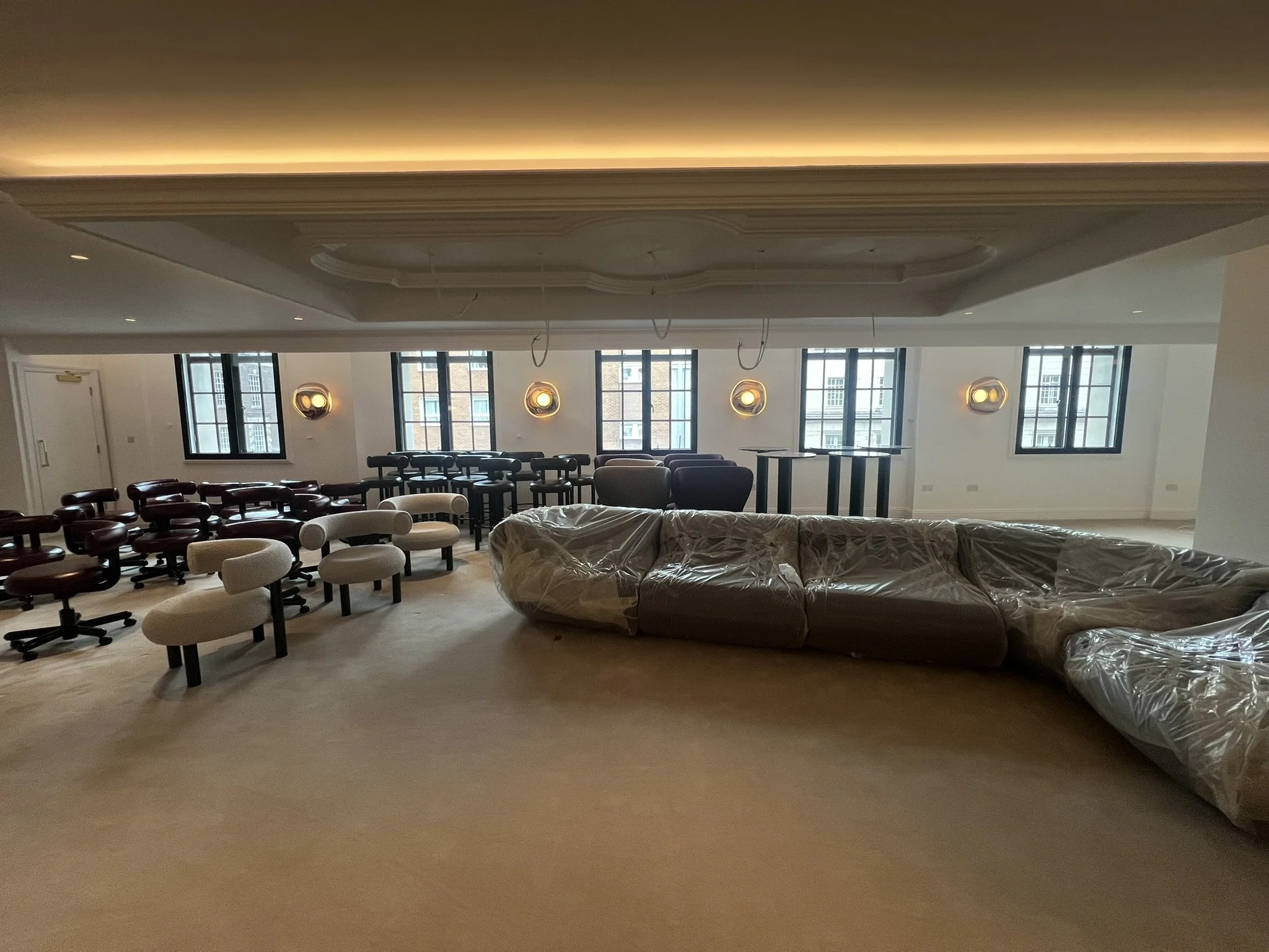Spacious lounge with sofas covered in plastic, upholstered chairs, café tables, large windows, and decorative wall lights.