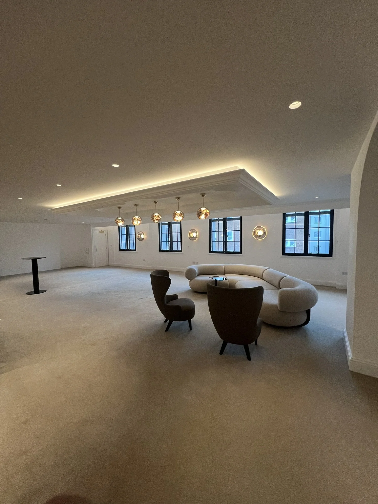 Modern, spacious lounge with a curved white sectional sofa, dark lounge chairs, round table, pendant globe lights, recessed lighting, and large black-framed windows.