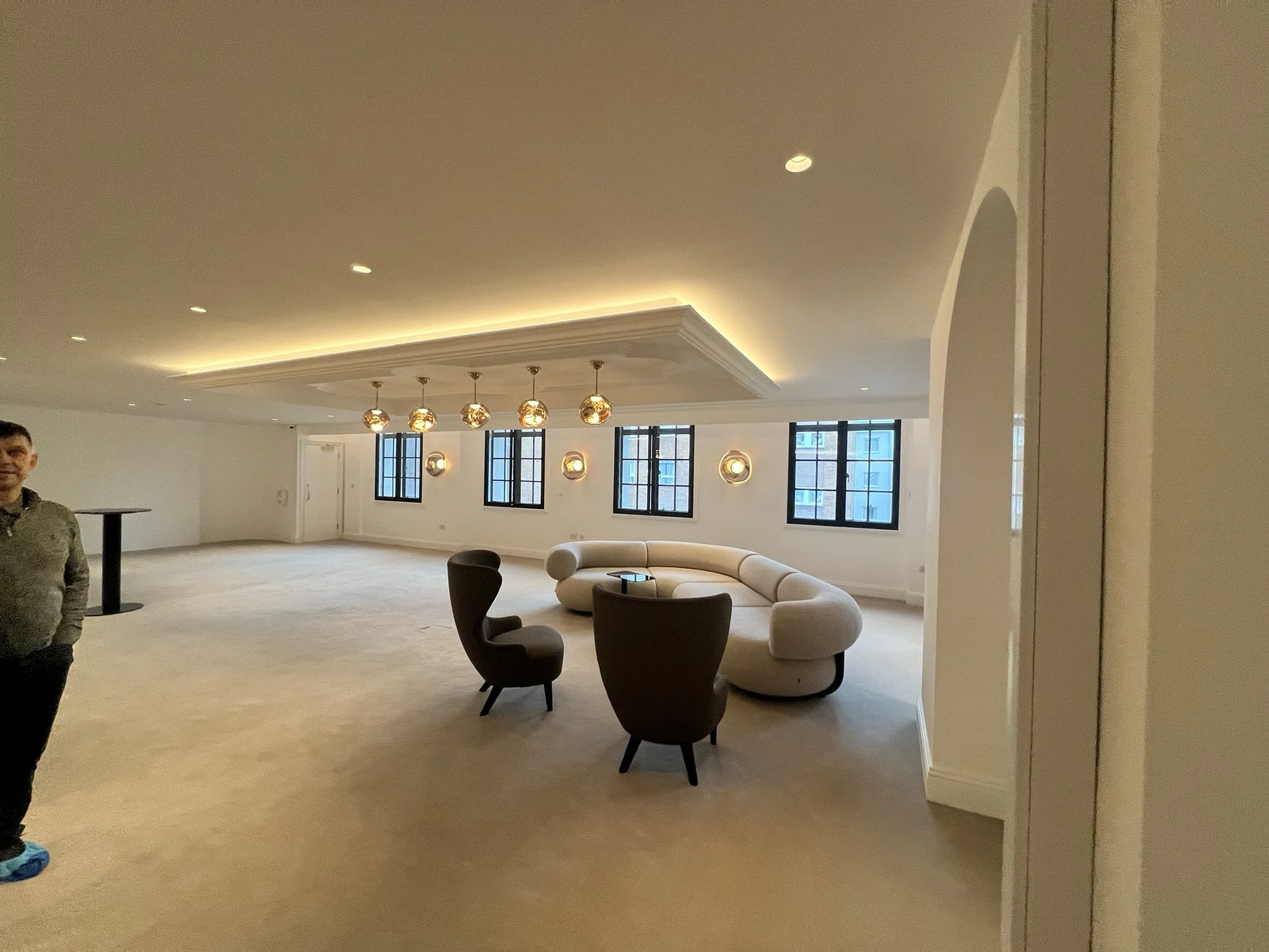 Spacious modern lounge with curved cream sofa, dark armchairs, pendant lights, large windows, and decorative ceiling molding