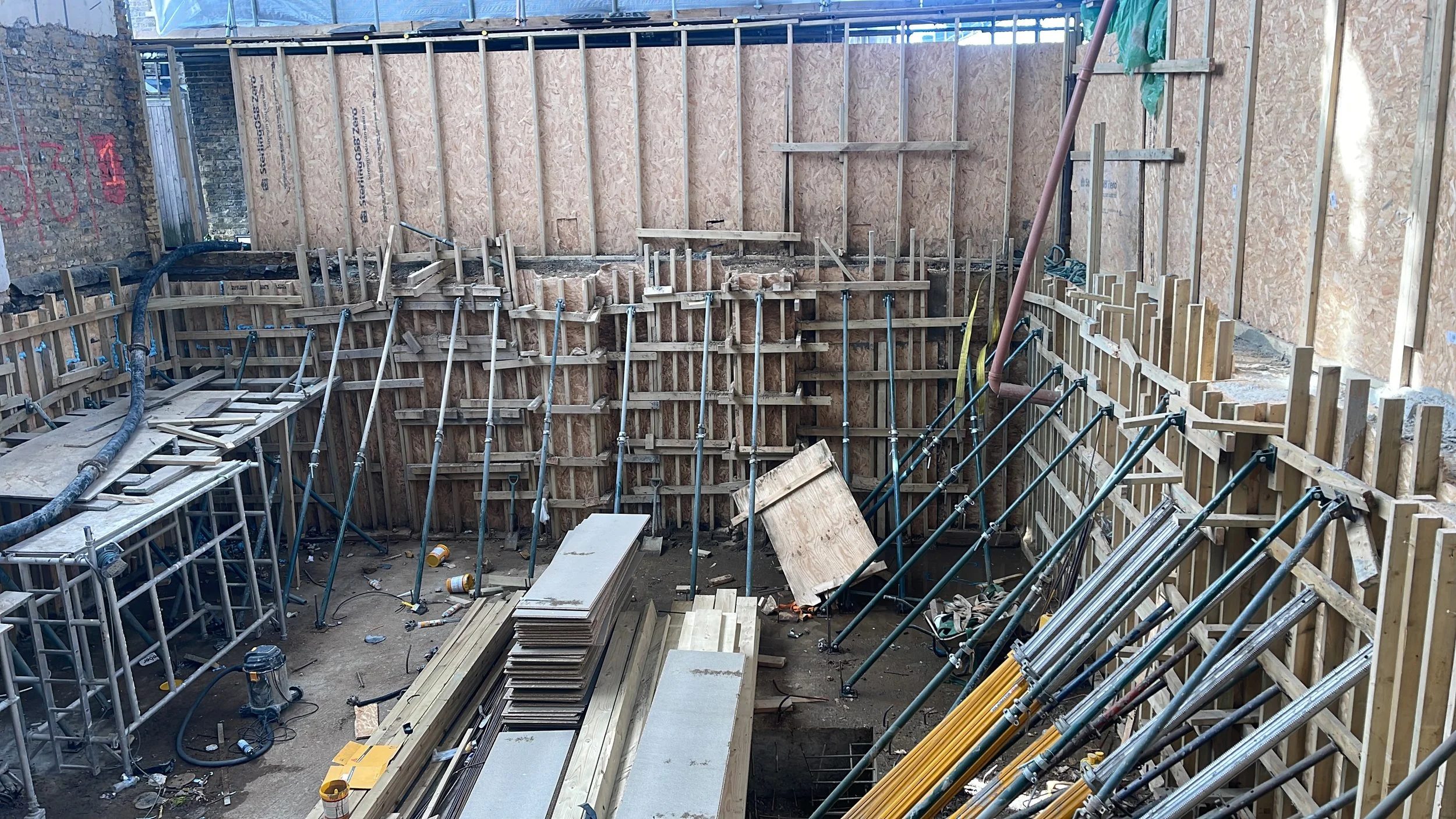 Construction site interior with plywood wall forms, metal support braces, scaffolding, lumber, and building materials