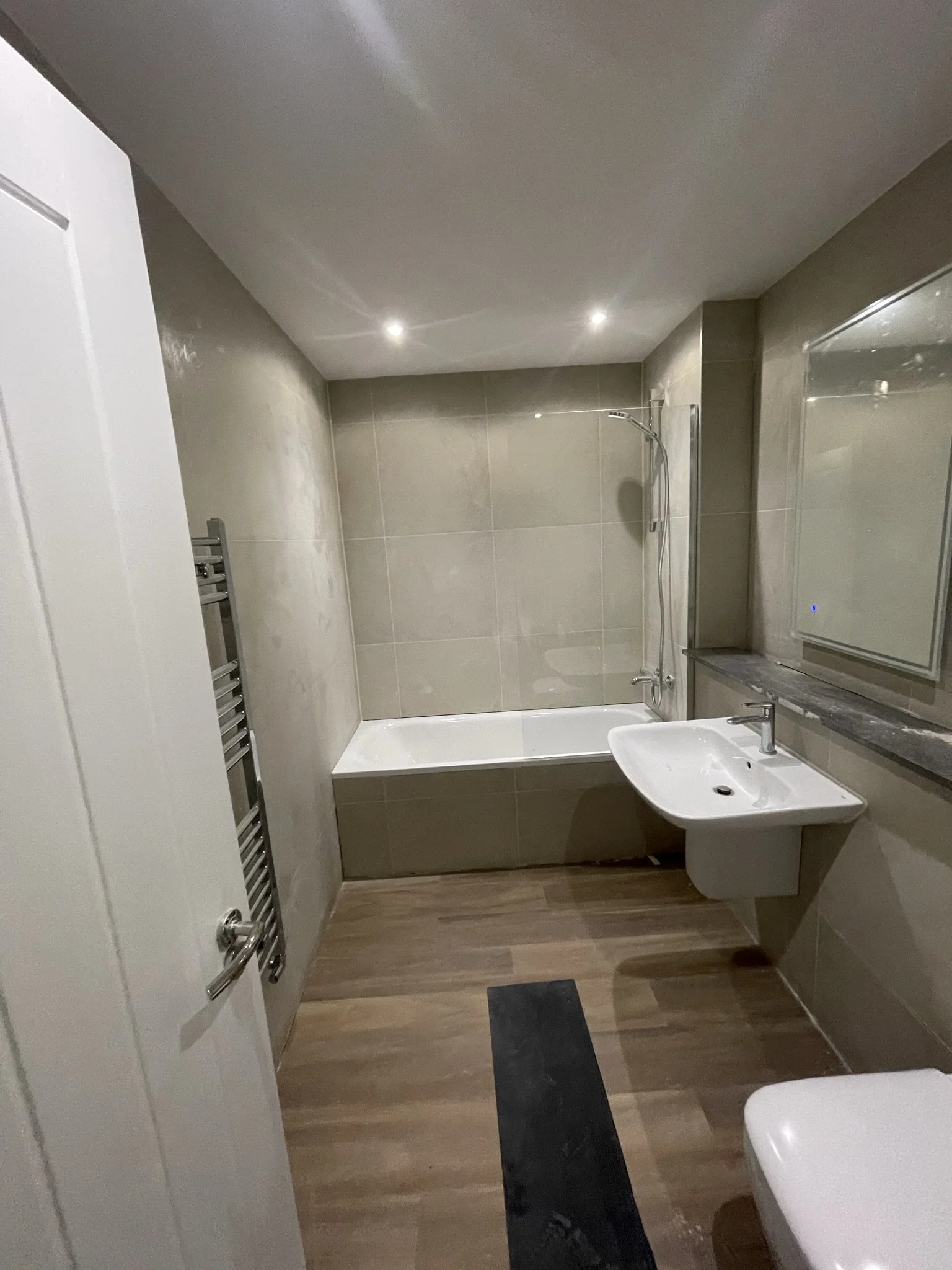 Modern beige-tiled bathroom with bathtub and shower, wall-mounted sink, heated towel rack, mirrored cabinet, and wood-look flooring