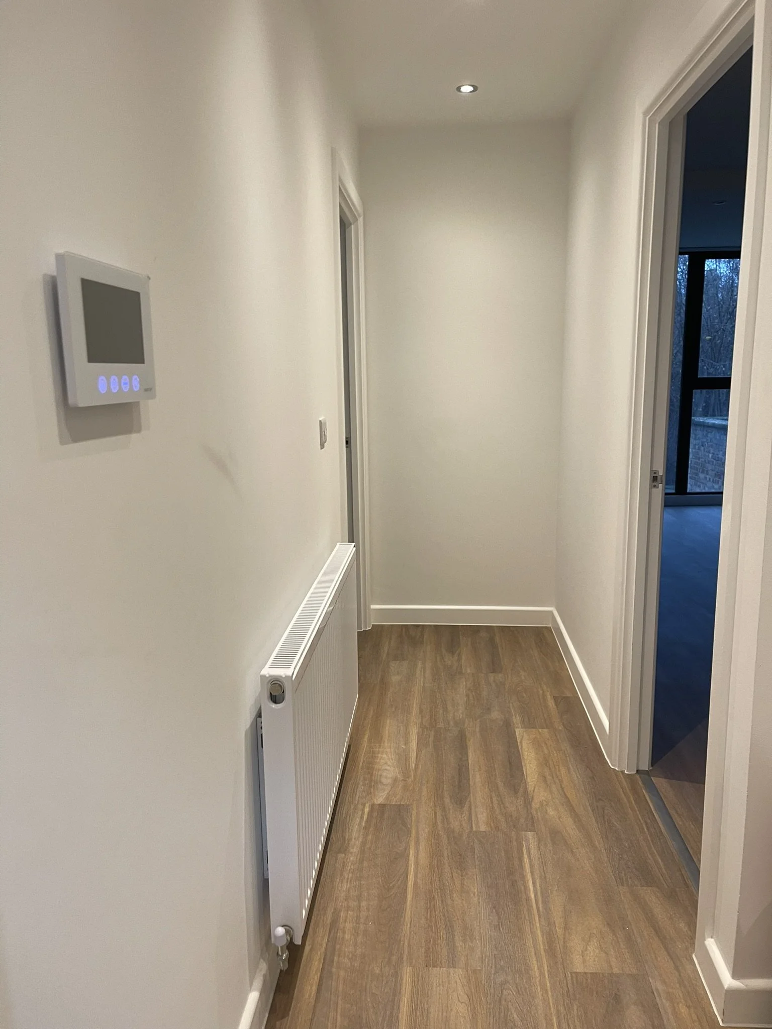 Bright residential hallway with wood-look flooring, white radiator, wall thermostat, recessed ceiling light, and open door to a room with large windows.