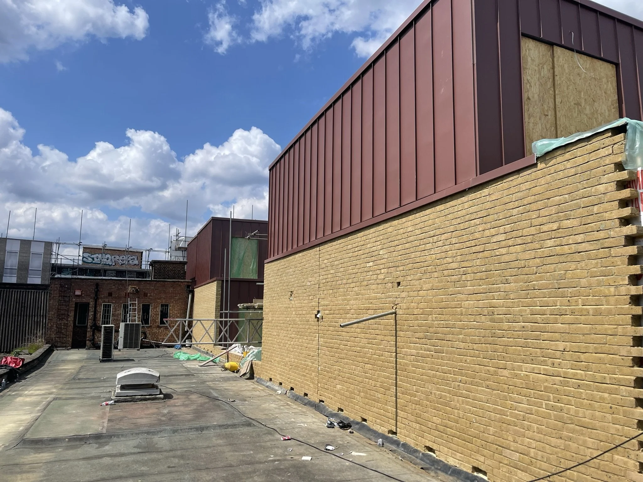 Commercial building rooftop under construction with yellow brick walls, burgundy metal cladding, scaffolding, and equipment beneath a blue sky
