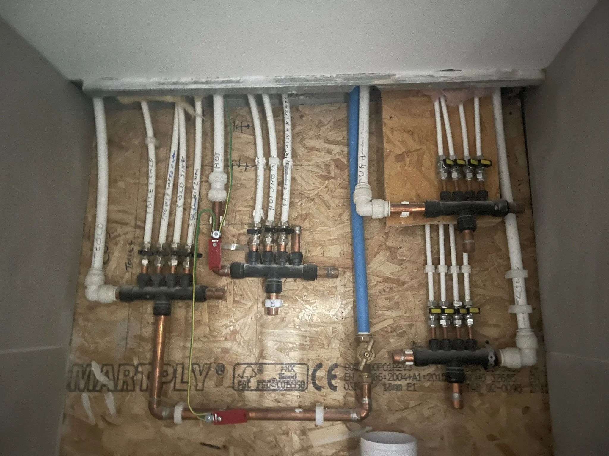Exposed residential plumbing manifold with labeled PEX lines, copper pipes, shutoff valves, and blue water supply tubing mounted on an OSB wall.