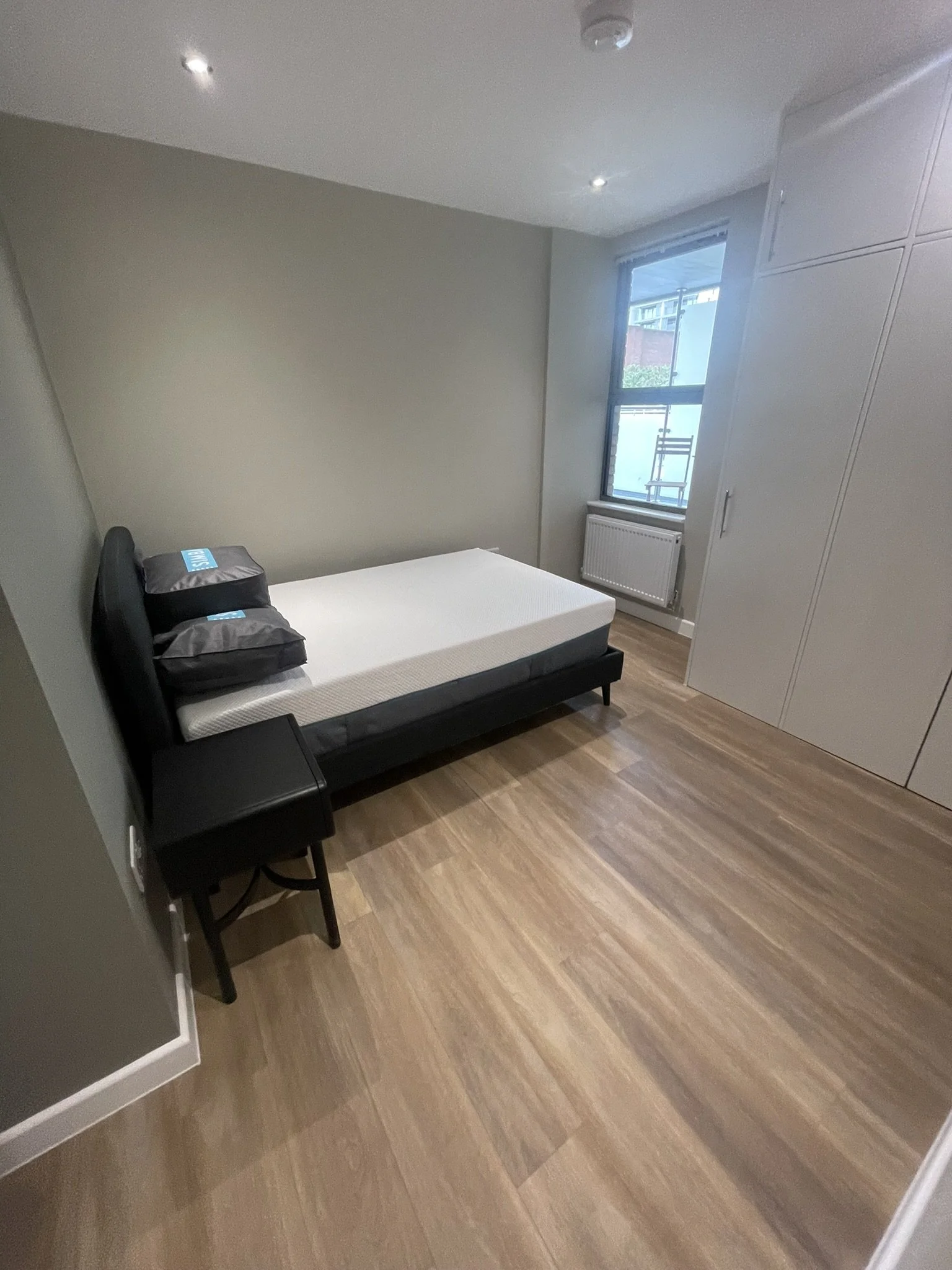Modern bedroom with a black bed, white mattress, pillows, hardwood flooring, window, radiator, and built-in wardrobe.
