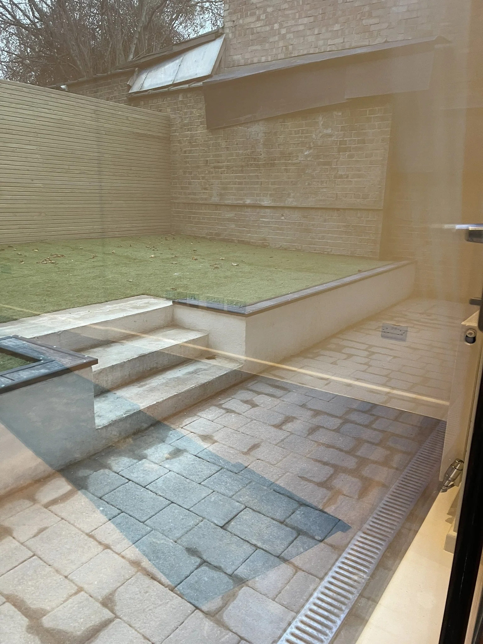 Brick courtyard with artificial grass, stone steps, paved patio, drainage channel, and surrounding walls viewed through a glass door