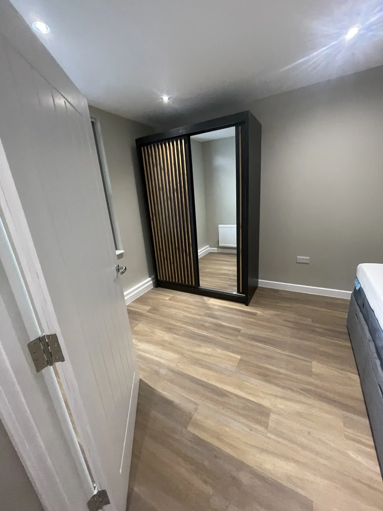 Modern bedroom with wood flooring, gray walls, recessed ceiling lights, a bed, and a black wardrobe with slatted wooden doors.