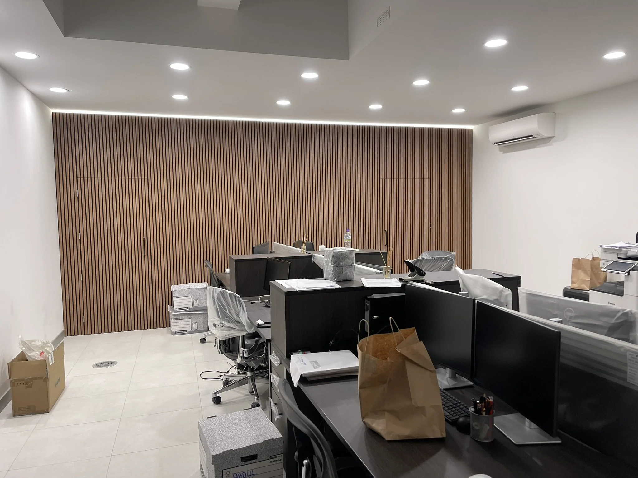 Modern office interior with black desks, computers, office chairs, wood slat accent wall, recessed lighting, and moving boxes