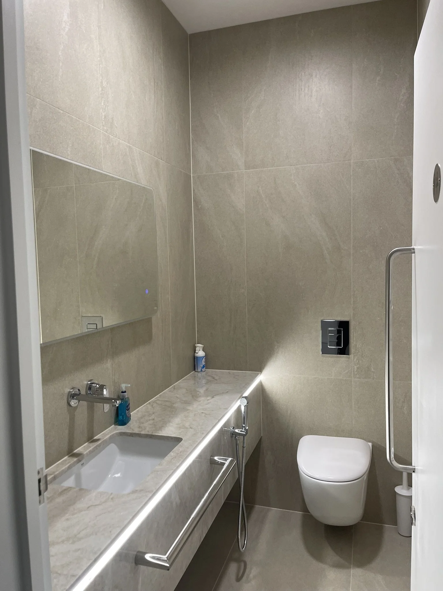 Modern beige-tiled bathroom with a bathtub, wall-mounted sink, toilet, large mirror, and chrome fixtures