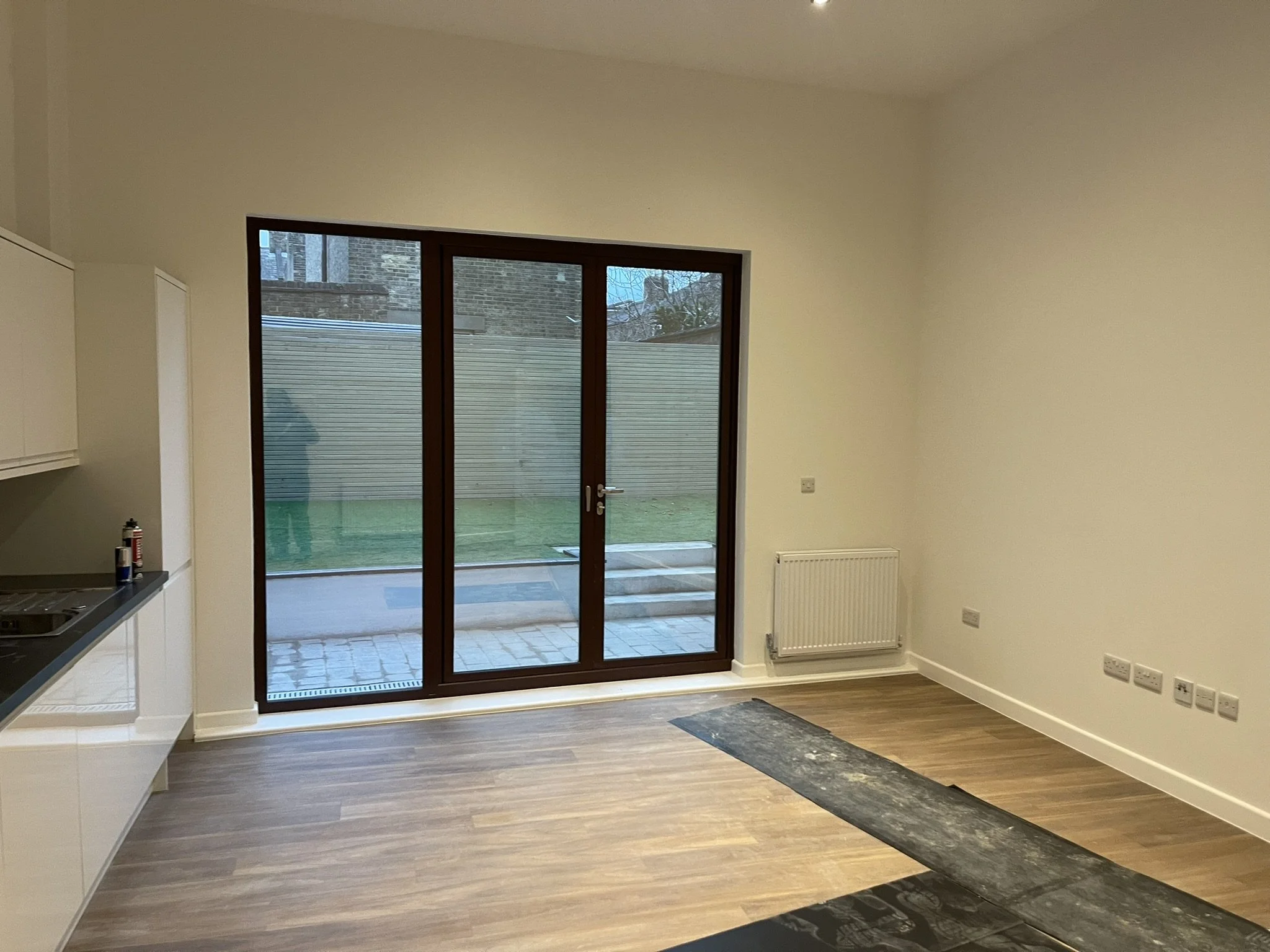 Modern open-plan kitchen and living area with wood flooring, dark-framed patio doors, white walls, and a wall-mounted radiator.