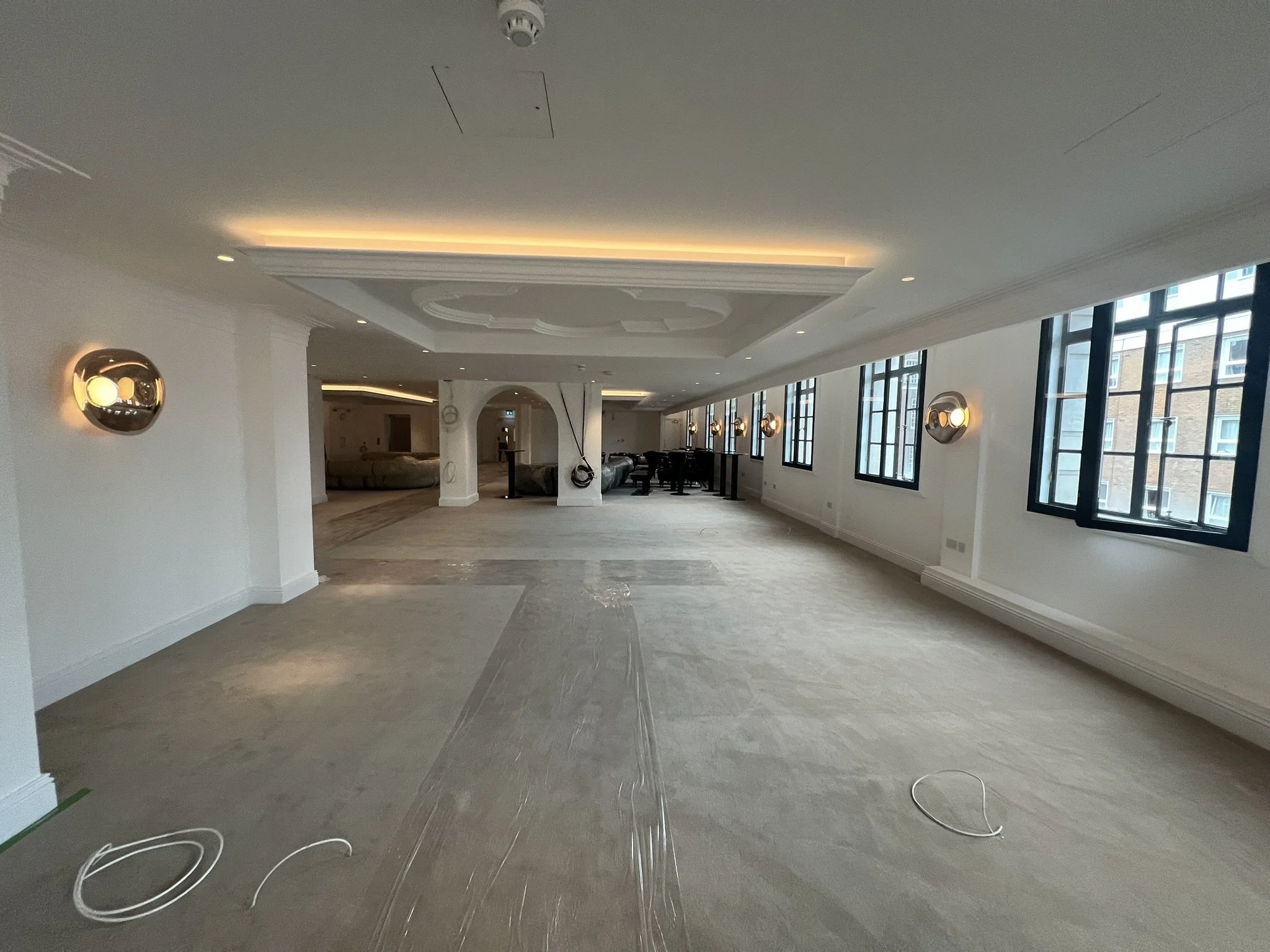 Spacious elegant interior room under renovation with ornate ceiling, wall sconces, large windows, lounge seating, and protective floor covering