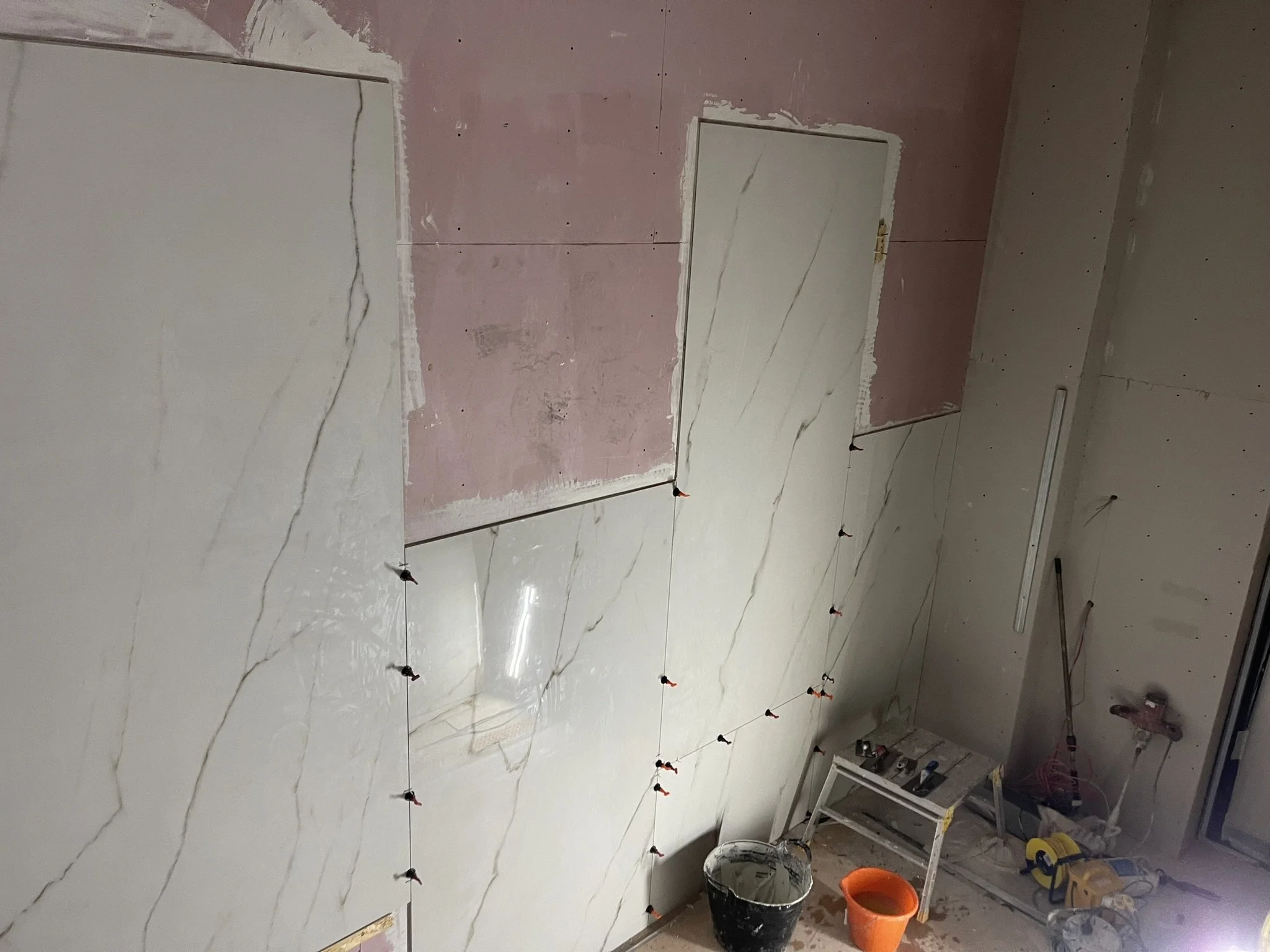 Bathroom renovation with large marble-look wall tiles, leveling clips, unfinished drywall, tools, and buckets.