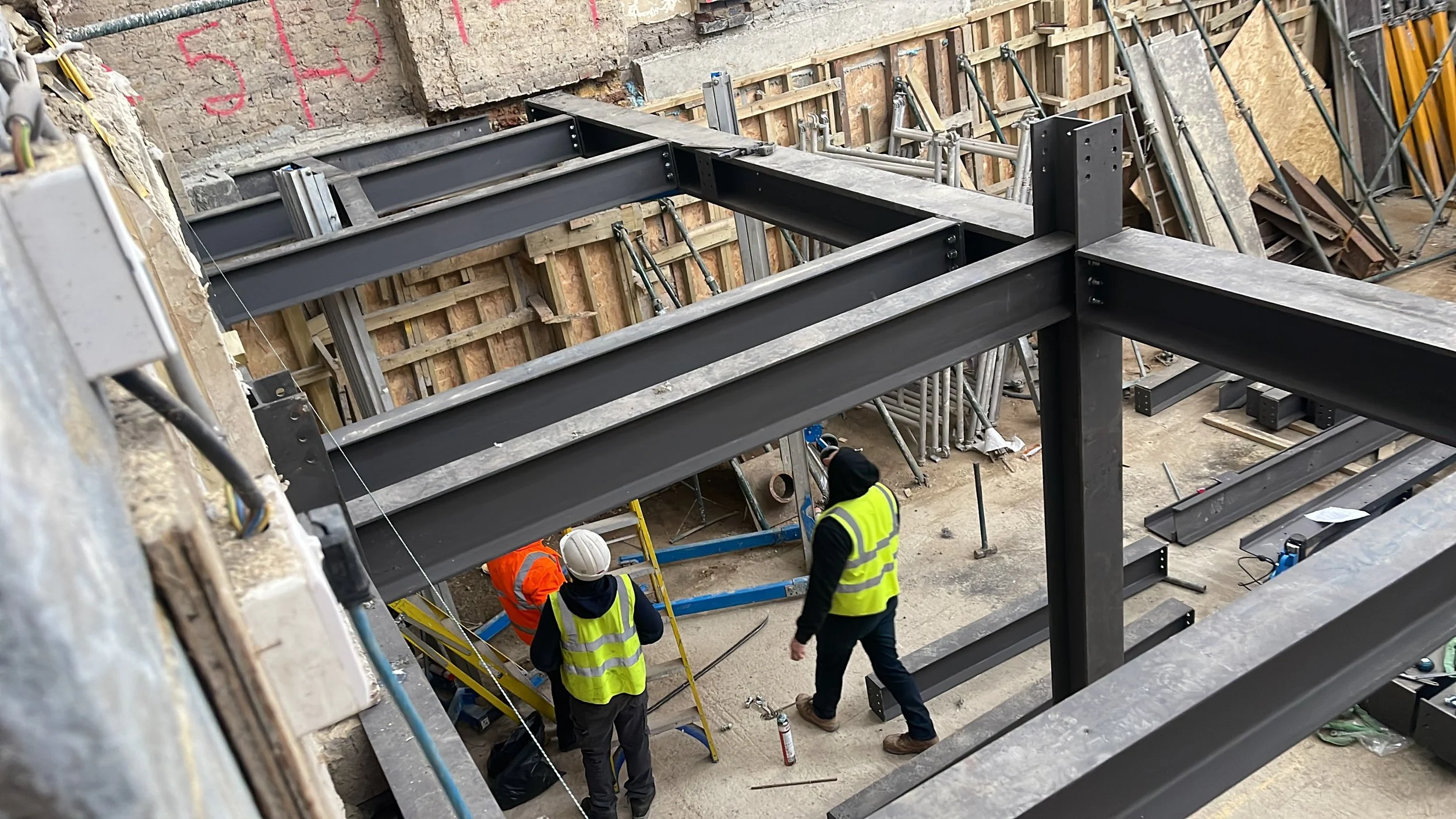 Construction workers in high-visibility vests installing structural steel beams inside a partially renovated building.