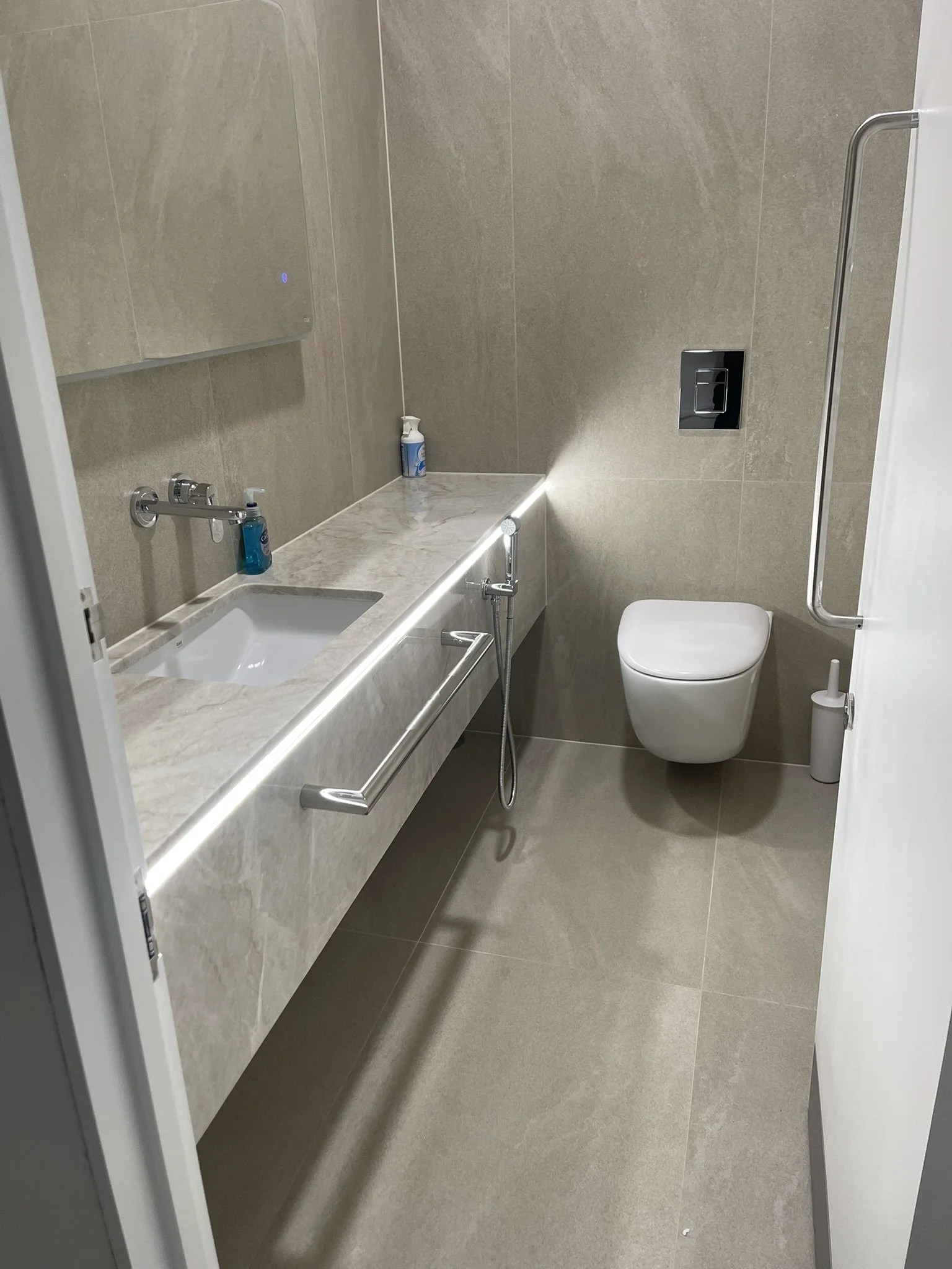 Modern beige-tiled bathroom with a bathtub, wall-mounted toilet, bidet sprayer, chrome grab bars, and illuminated mirror