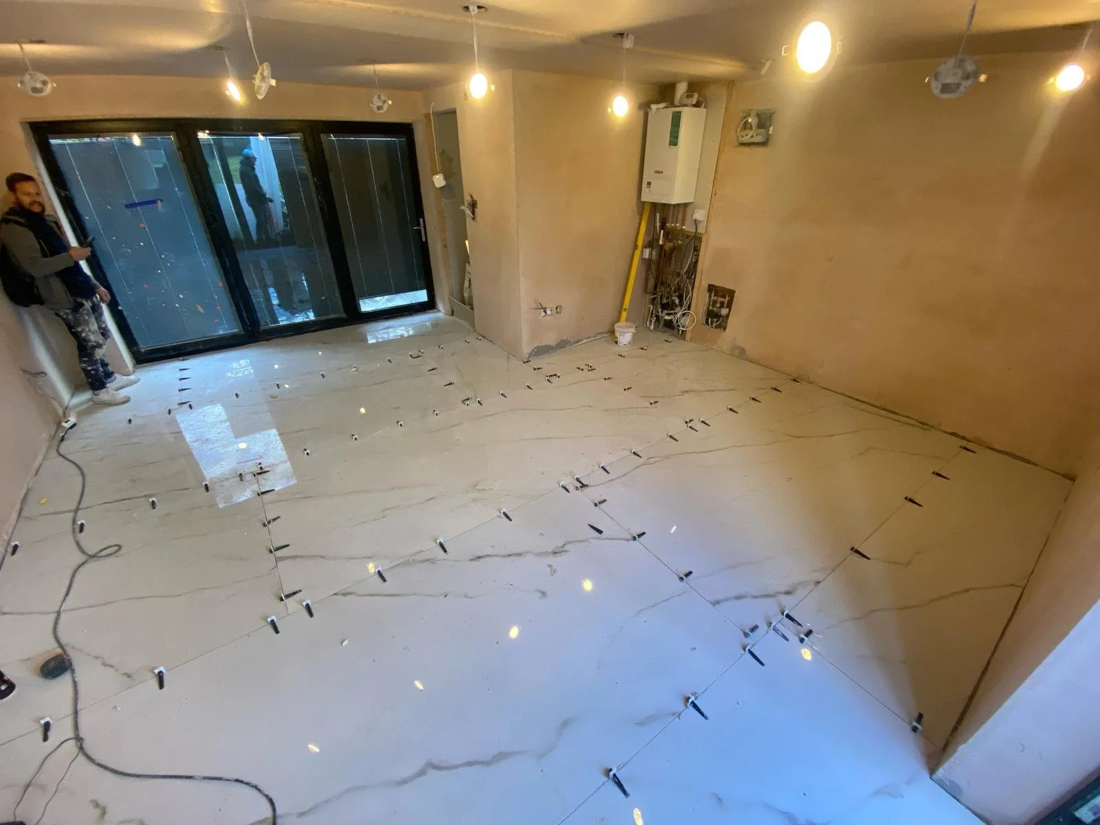 Renovated room with large white marble-look floor tiles, leveling spacers, exposed plumbing, and a worker by the glass doors