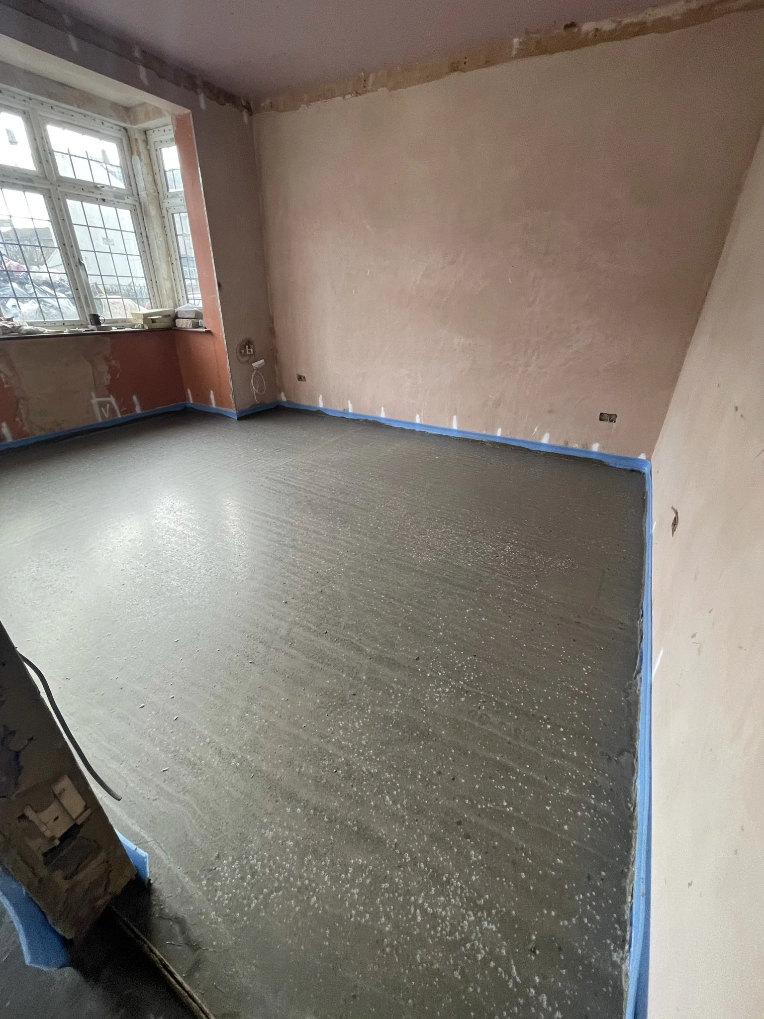 Unfinished room under renovation with a freshly poured concrete floor, exposed wall edges, and blue painter’s tape along the baseboards